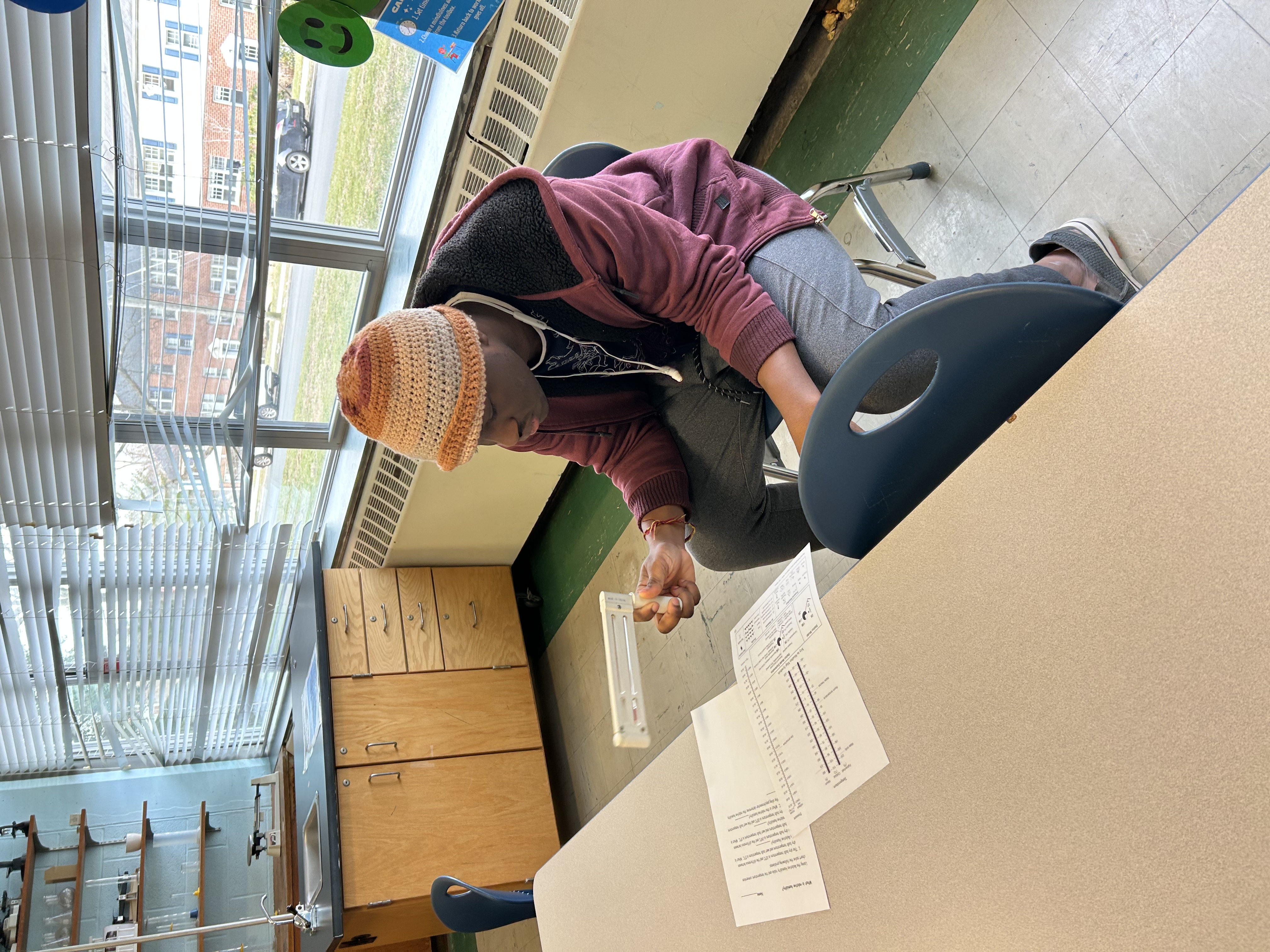 A student sits at a table in the classroom holding a psychrometer