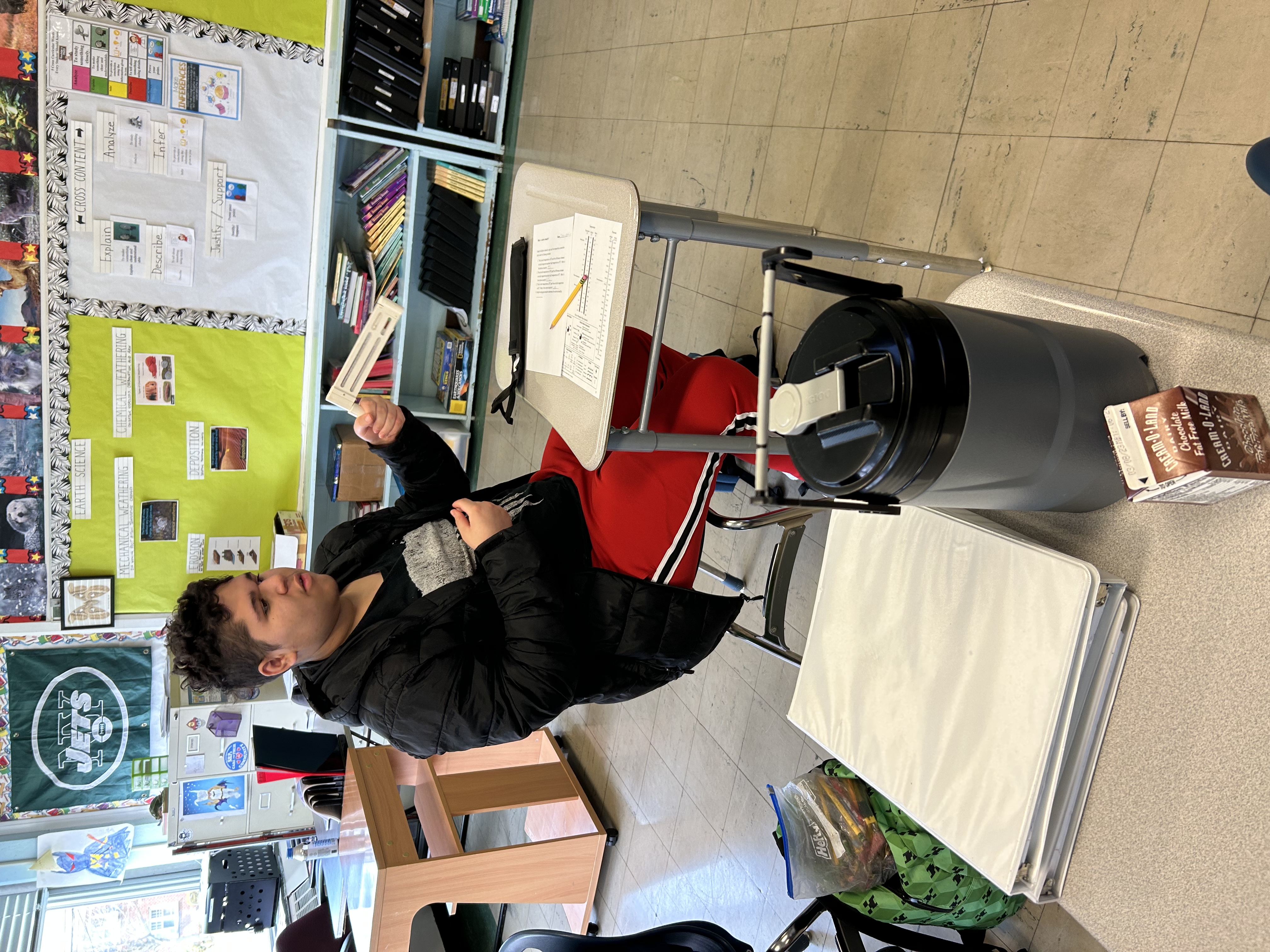 A student sits at his desk holding a science tool called a psychrometer