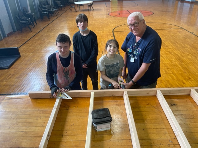 three students pose with Mr. Babiak in front of their wooden project which lays on stage in the gym