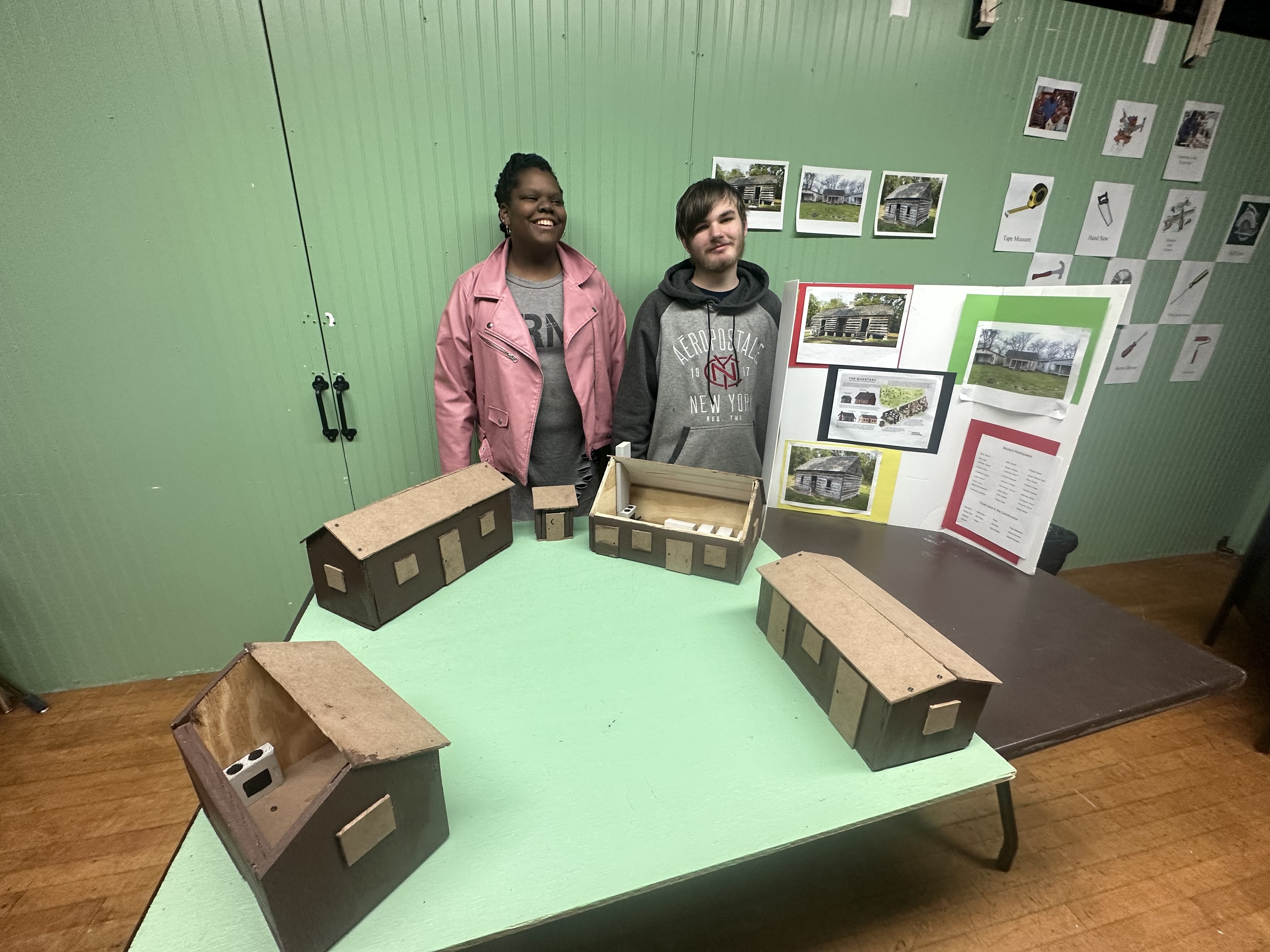 Two students pose behind the wooden model project they made. The project is wooden houses representing houses which slaves lived in. Their tri-fold poster board with information on it is to the side of them