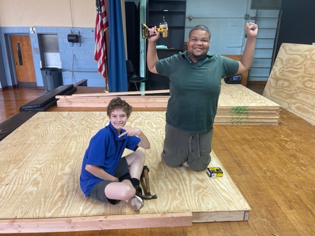 Students pose in class holding tools they are using to create their wooden project. One is sitting giving the peace sign and the other one is kneeling with his arms raised