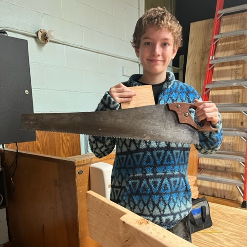 A student stands smiling holding a saw and some wood in class