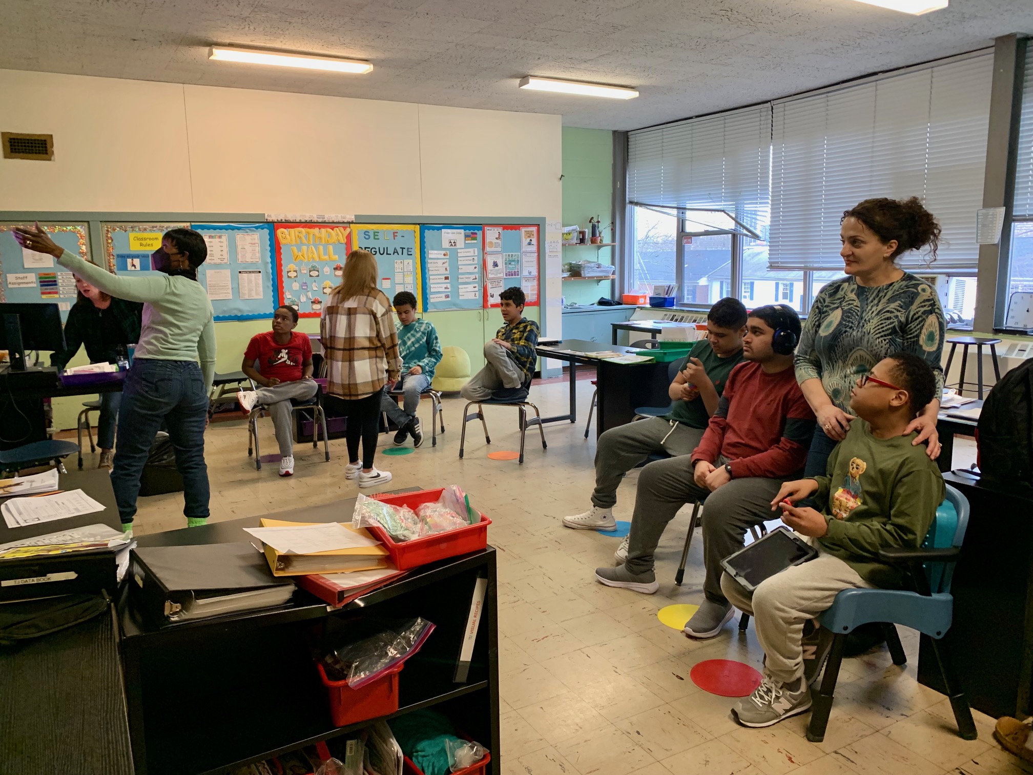 Students sit in a semi-circle in class ready for group while their aides help them