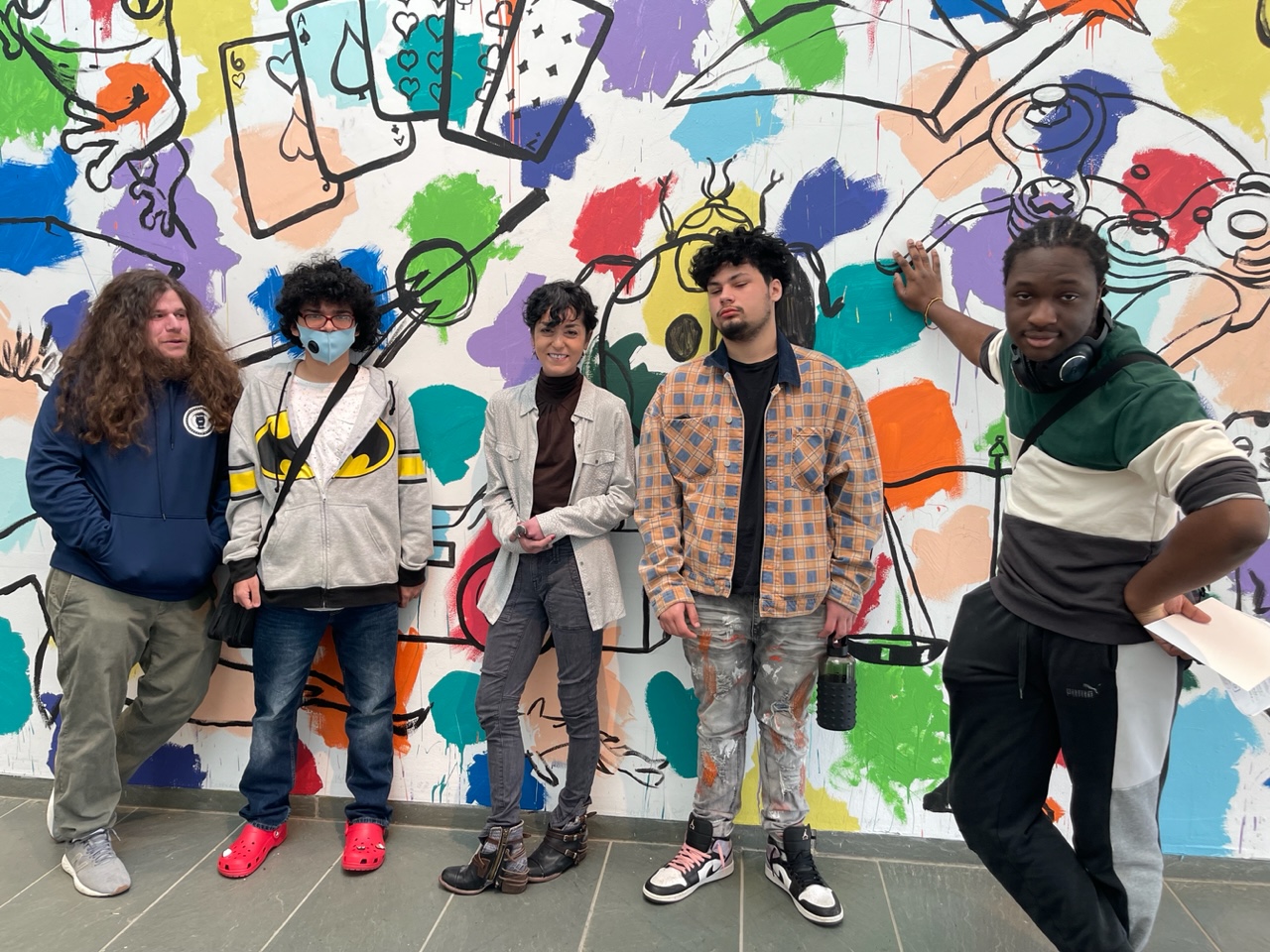 Students stand for a group shot with the art teacher Ms. Mossa and aide Mr. Rubin in front of a colorful art mural which is different colored paint splotches on a white background