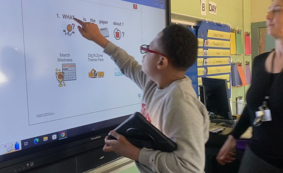 A student points to his work on the smart board while holding his talking device