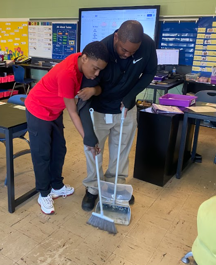 A staff member helps a student sweep the floor in the classroom by guiding the student's hands holding the broom