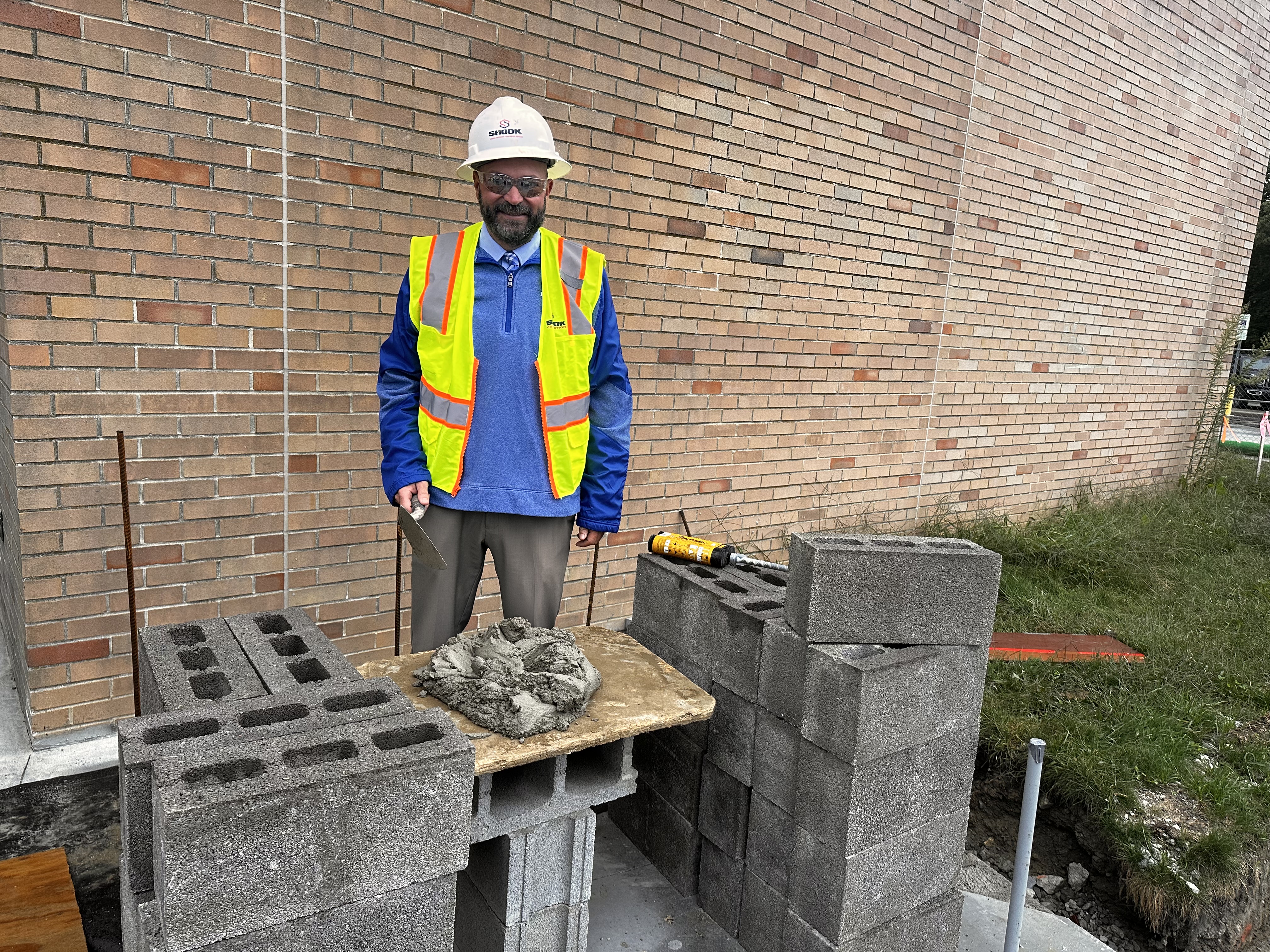 A man in a hard hat and yellow safety vest is standing with a trowel and cement on a construction site.