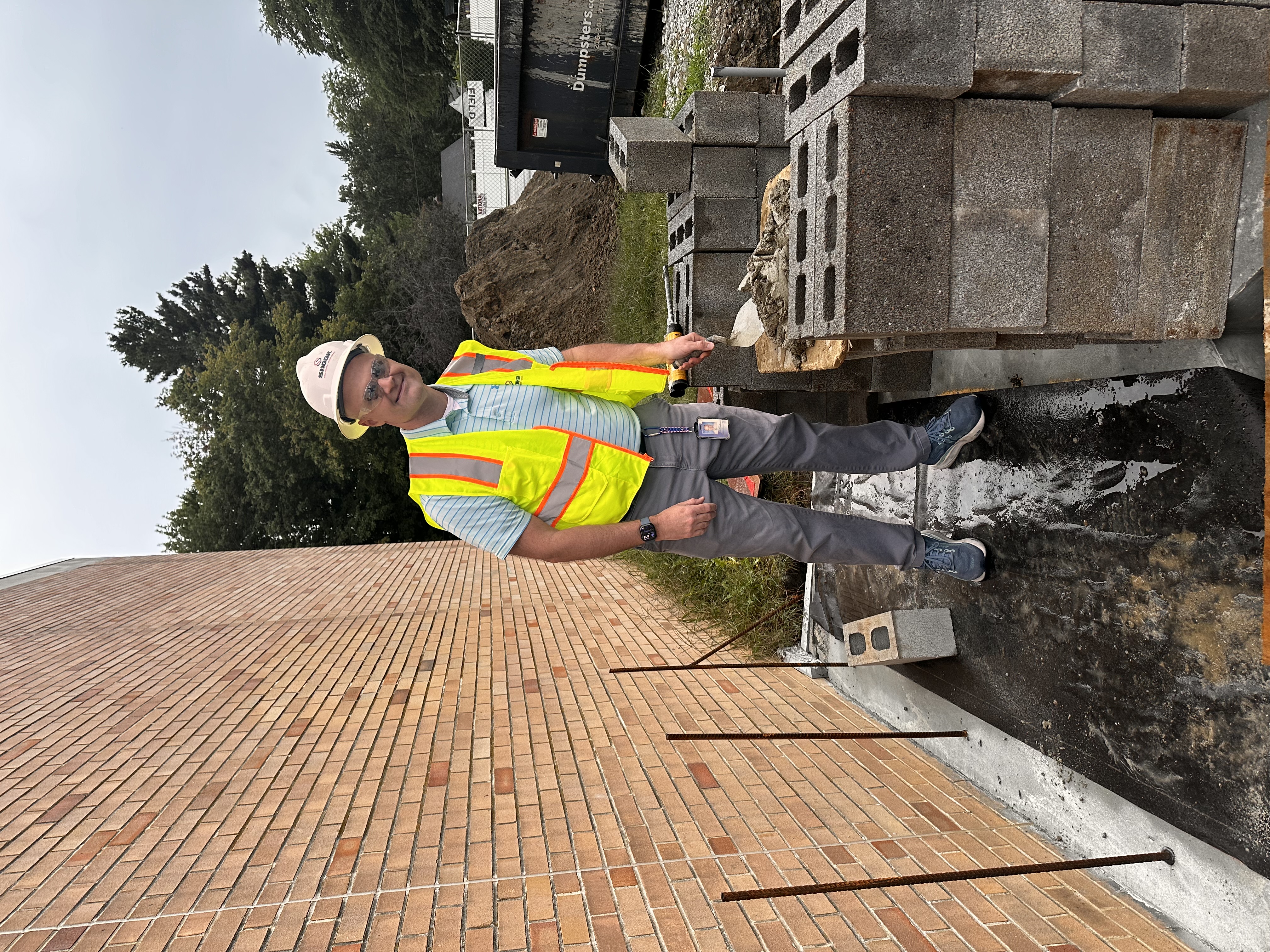 A man in a hard hat and yellow safety vest is standing with a trowel and cement on a construction site.