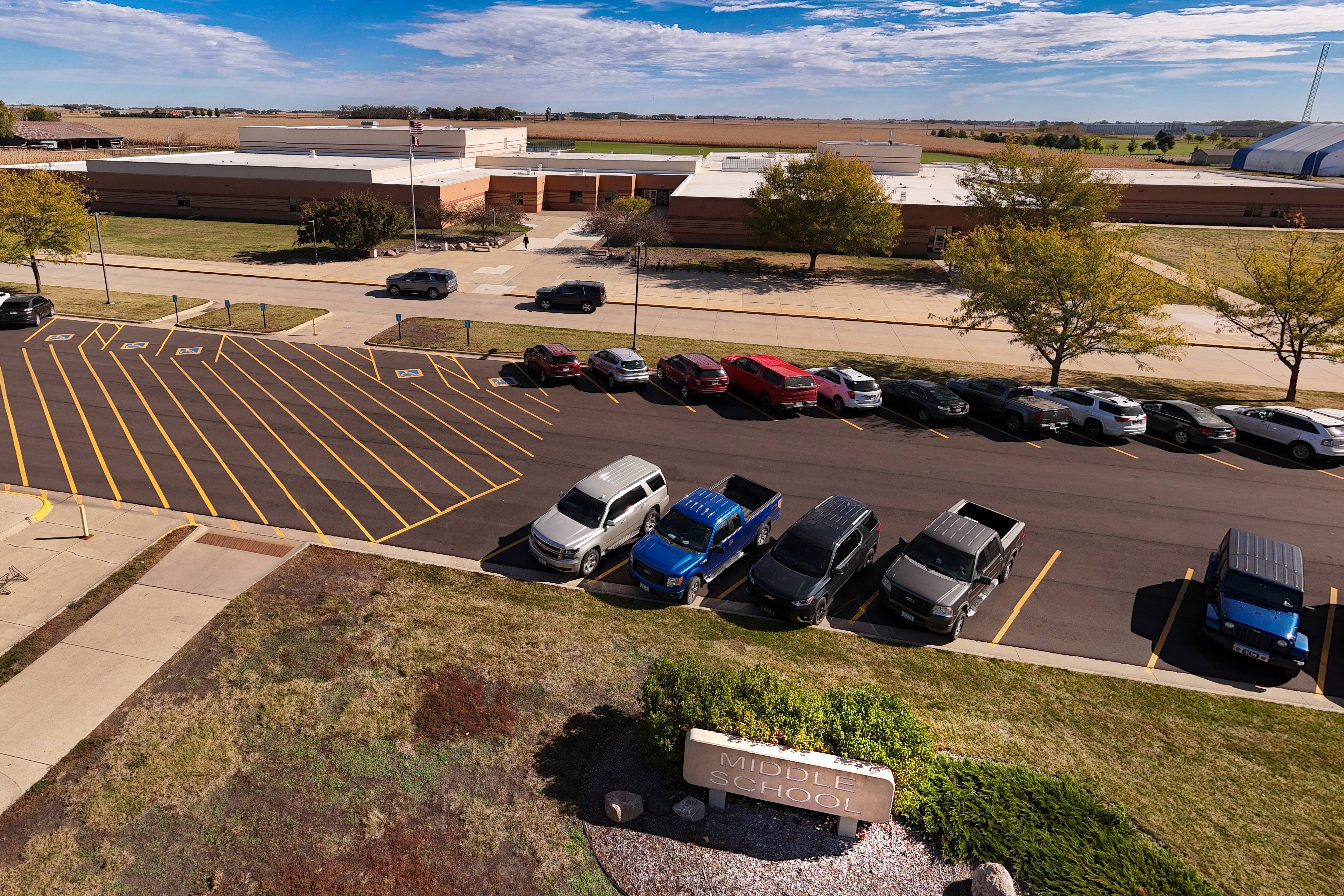 Aerial view of a parking lot with various cars parked. Building in background with greenery and open fields.