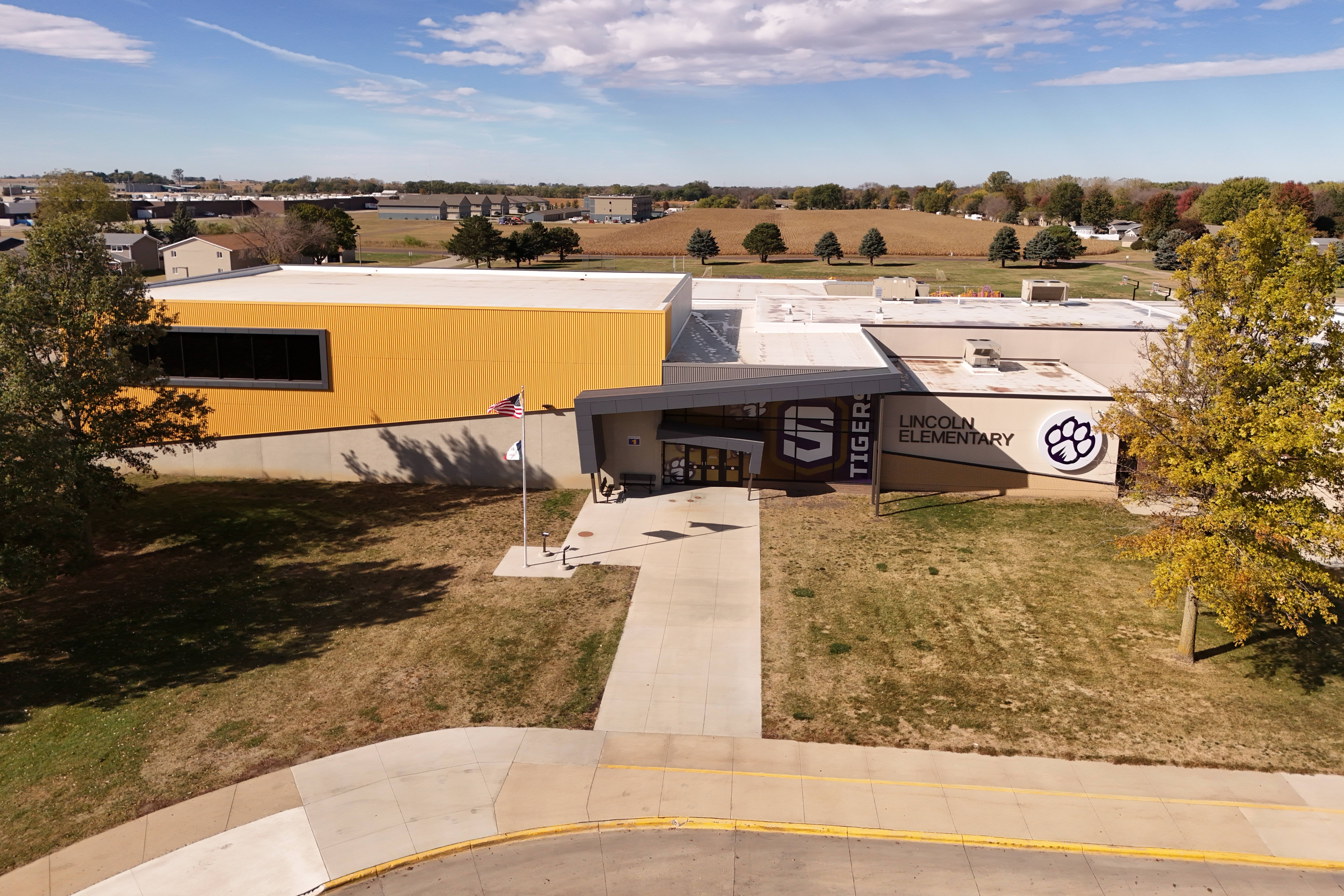 Aerial view of a yellow and white building with "S" logo and emblem, featuring a pathway, grassy area, and trees.