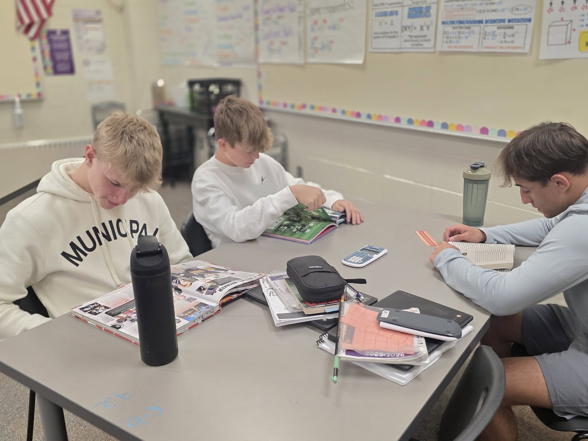 Three students sit around a table with books, a water bottle, and a tablet. Behind them, papers are pinned to a wall.