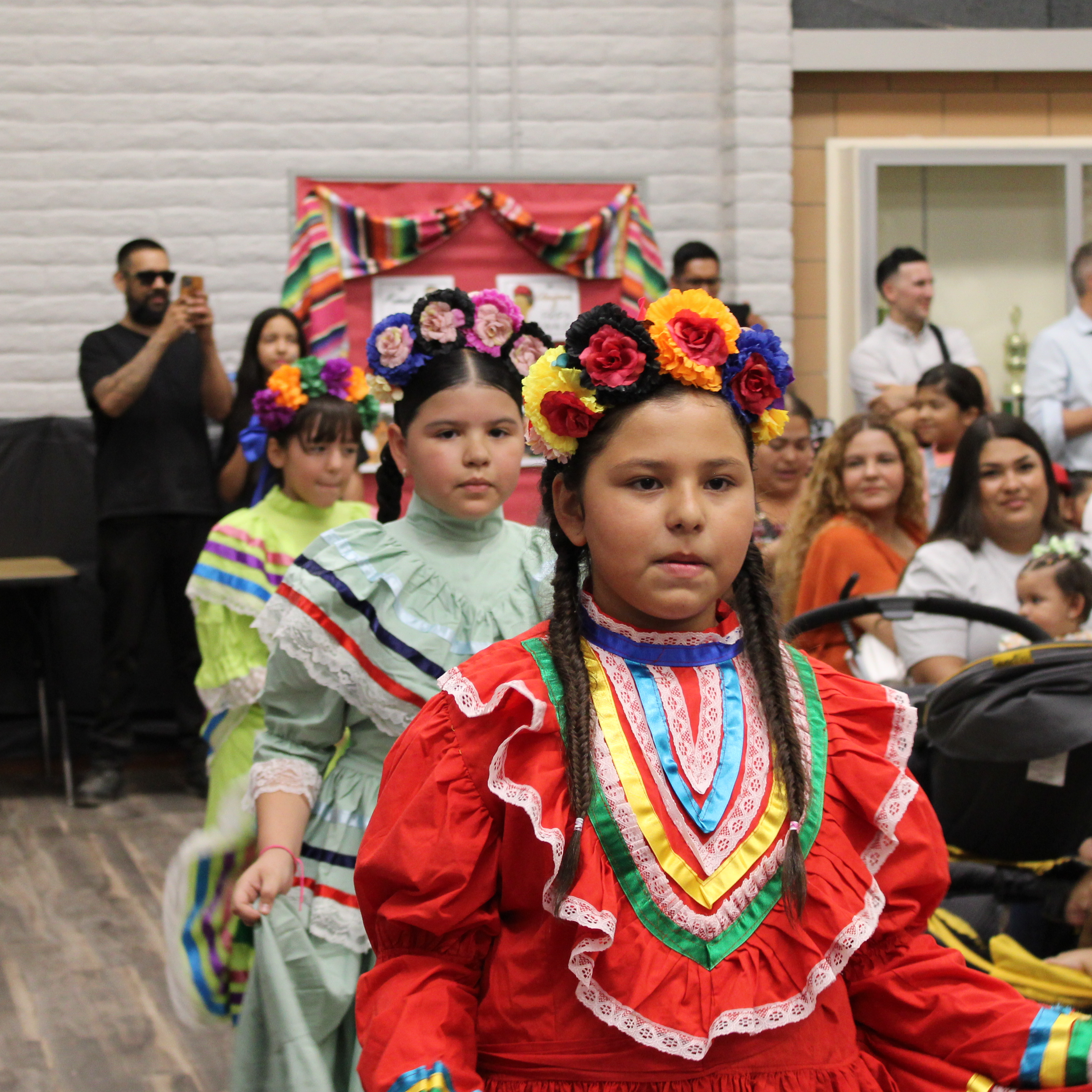 three elementary students dressed in cultural hispanic attire perform in front of crowd of parents  