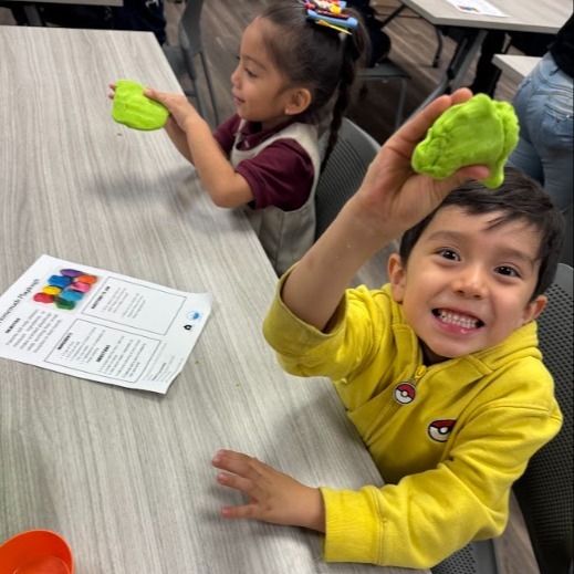 two elementary students sitting at table woking with play-dough. the student closest to the camera wearing a bright yellow jacket, enthusiastically displays his green play-dough to the camera 