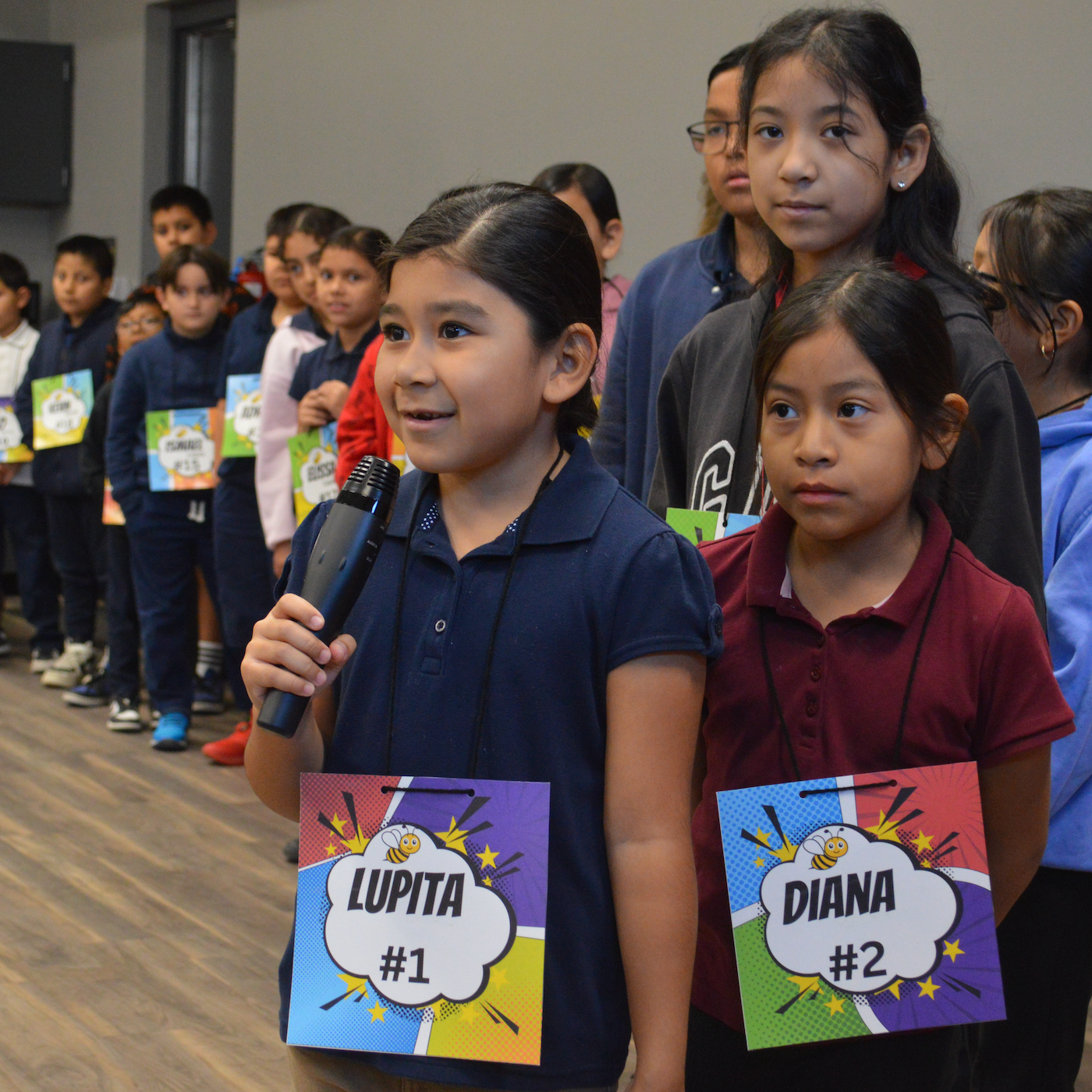 a young elementary student holding a microphone and spelling a word during school spelling bee. behind her is a long line of elementary students waiting for their turn at the front 