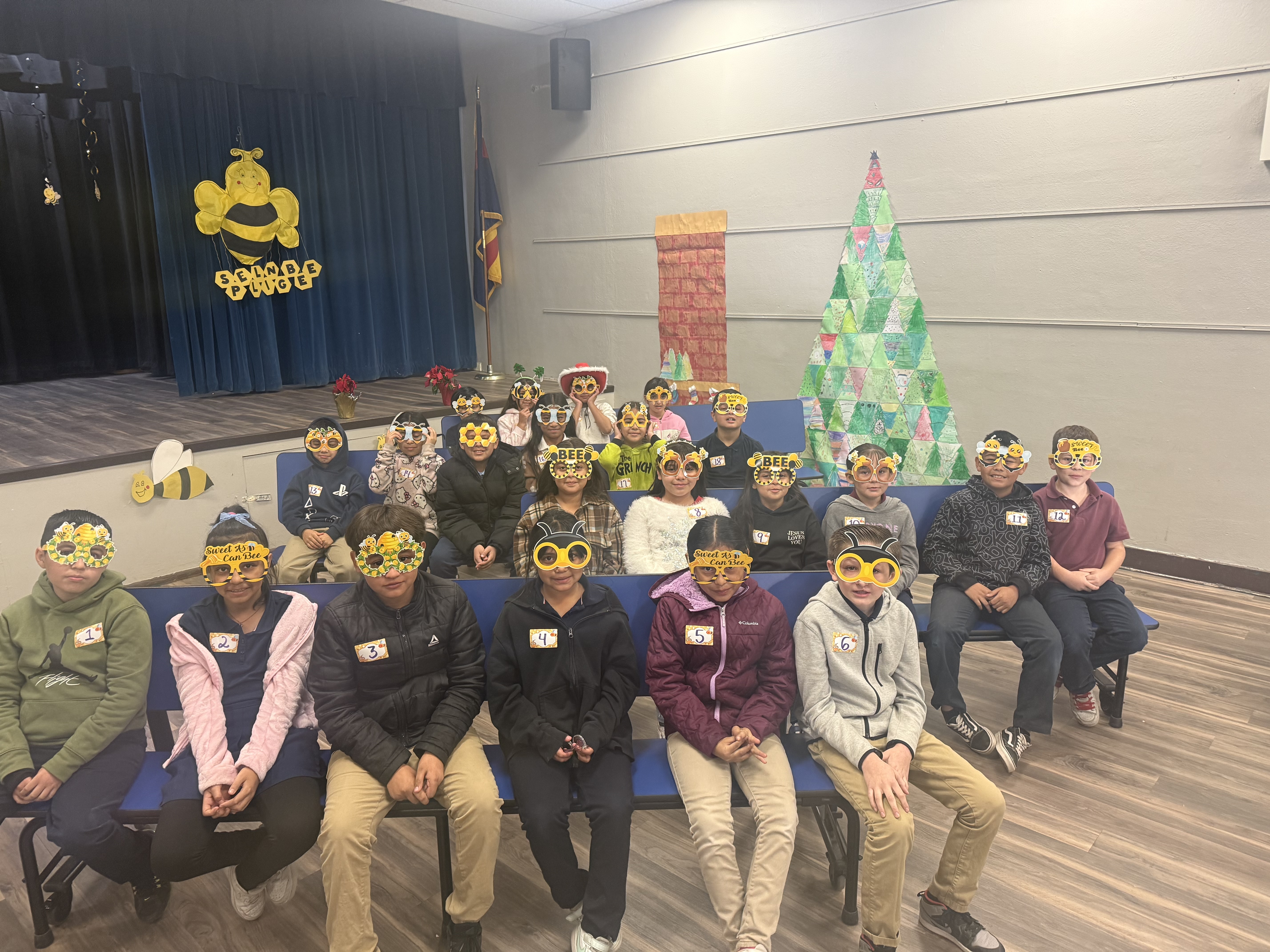 This image shows a group of children sitting together in the cafeteria. The children are wearing bee-themed masks and name tags, for the Spelling Bee. The background includes decorations such as a bee cutout with "Bee Kind, Bee Yourself" text. 