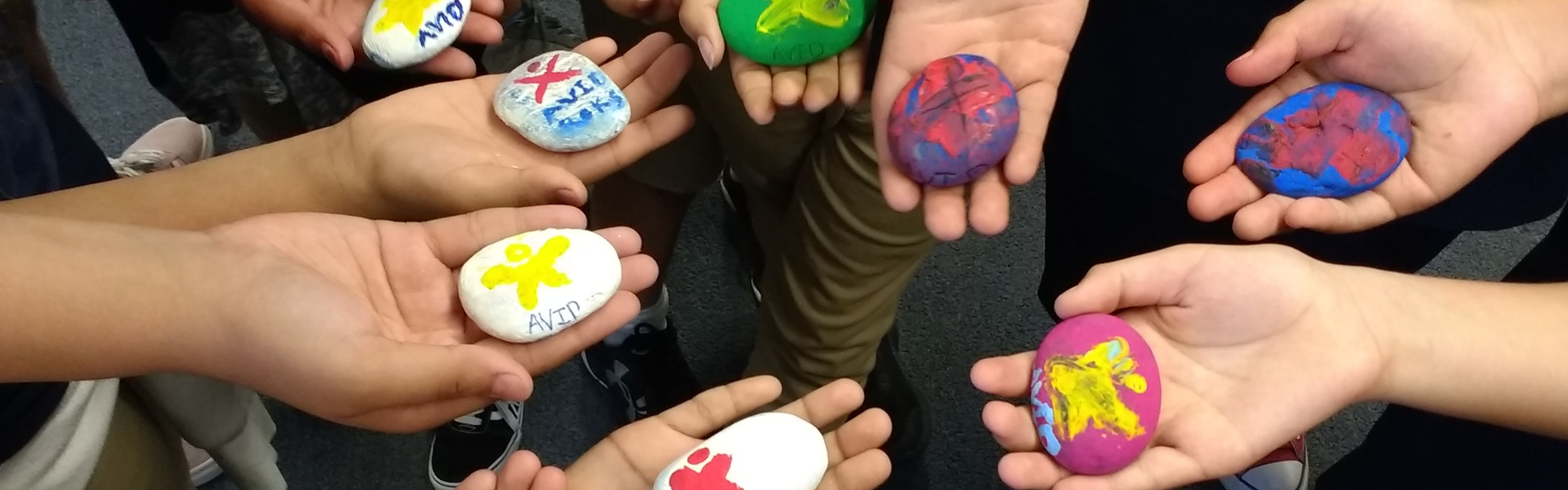 Multiple hands holding colorful painted rocks with various designs and patterns. The setting conveys creativity and a sense of community.