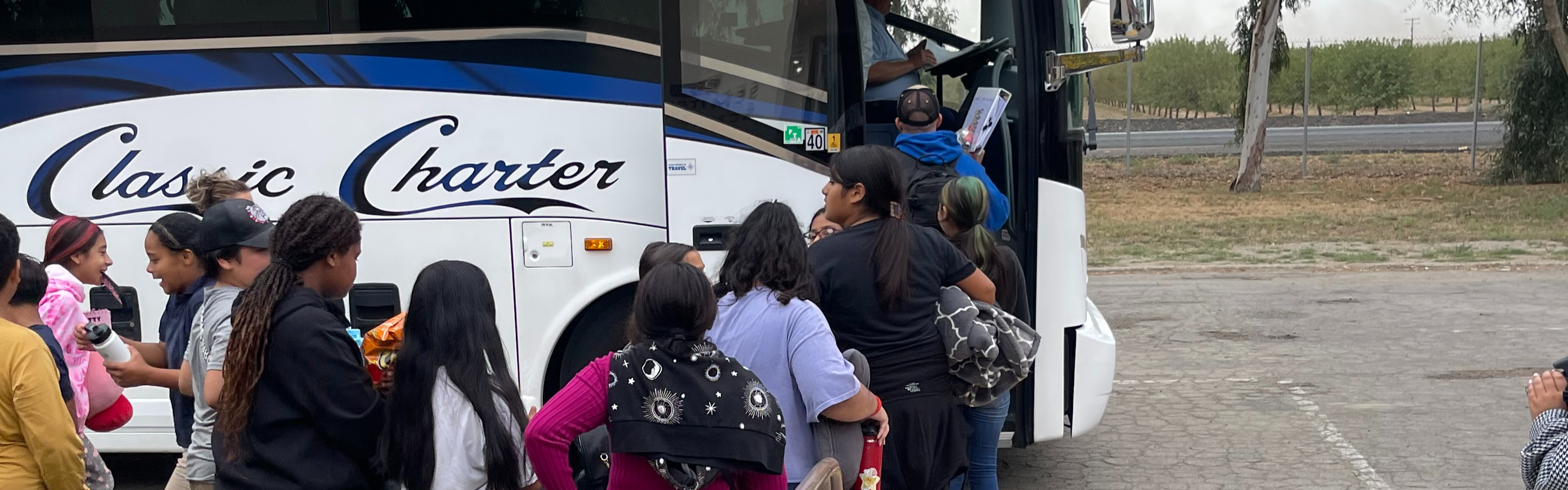 A group of people, mostly women, and children, are lined up to board a "Classic Charter" bus parked in a rural area, creating a sense of anticipation.