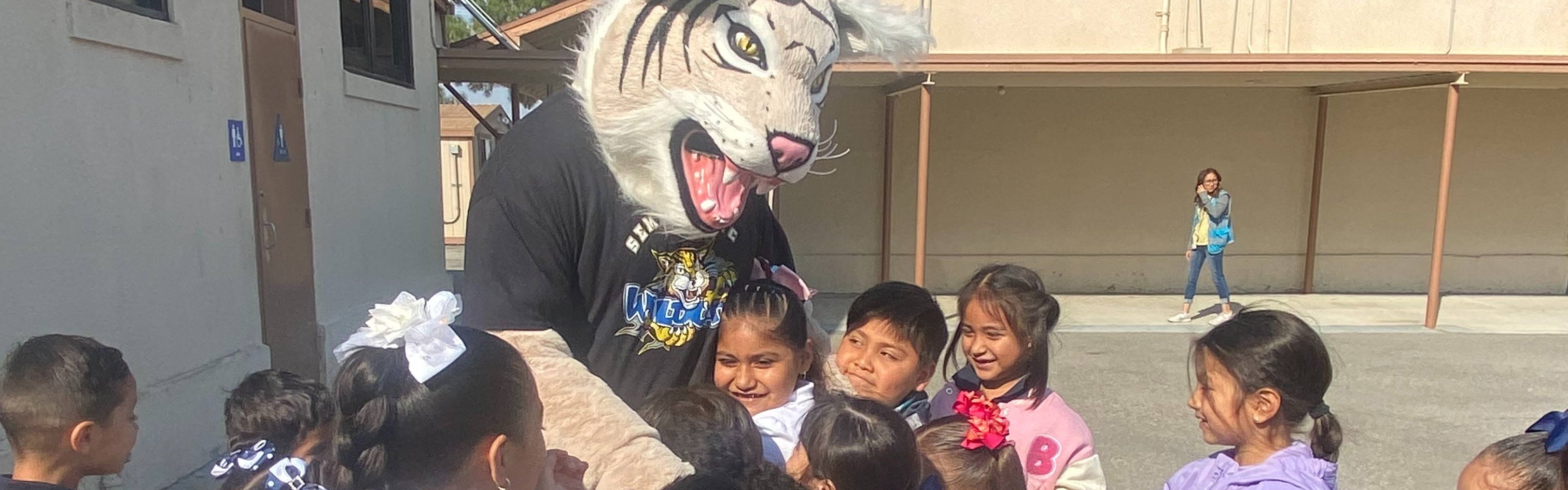 A person in a tiger mascot costume interacts playfully with a group of smiling children outdoors. The mood is joyful and engaging.