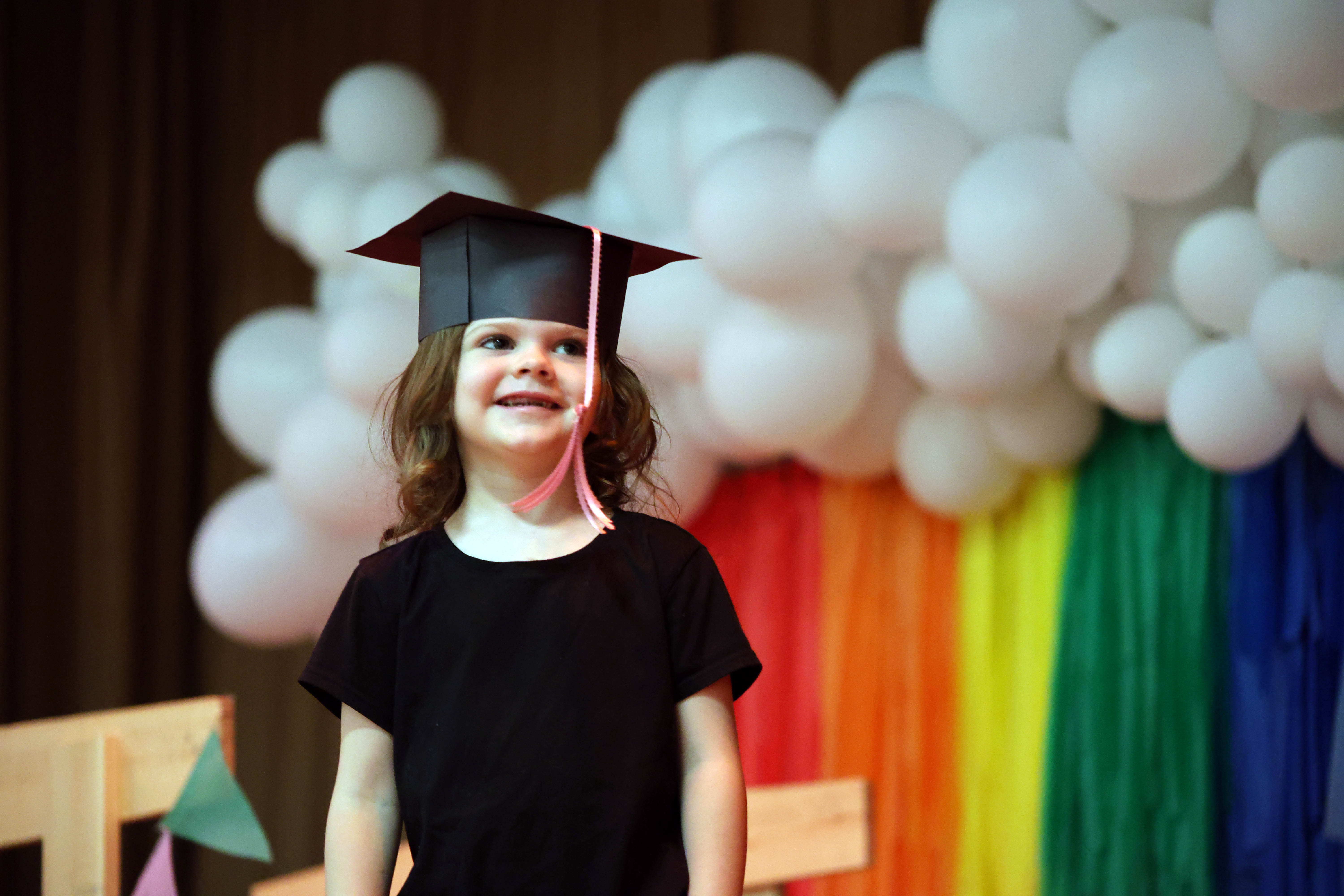 young girl in graduation cap smiling with rainbow and clouds stage set behind her