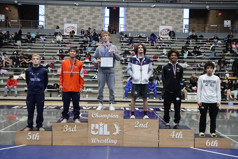 Competitors stand on a stage with podiums labeled 1st to 6th. One holds a medal. Audience in bleachers.