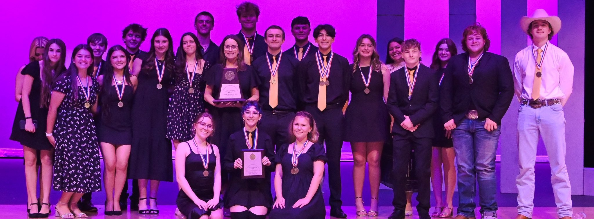 A group of people wearing black and medals, some seated, some standing, on a stage with a purple backdrop.