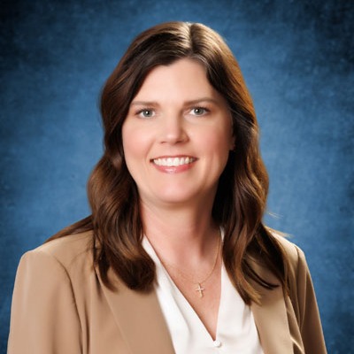 Woman with brown hair and a smile stands against a blue background. Wearing a white shirt and a brown blazer, she has a necklace.