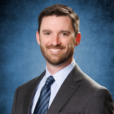 Man wearing a business suit and blue tie smiles at the camera. The background is a solid blue color.
