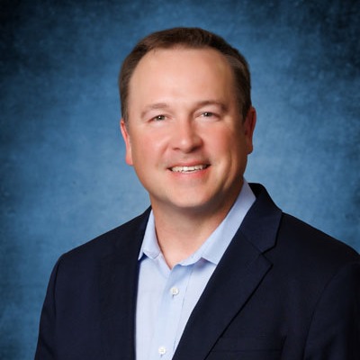 Man with short hair in a navy blue blazer and light blue shirt, smiling against a blue background.