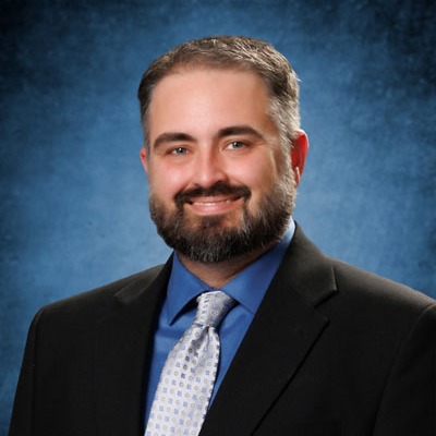 Man in suit and tie, smiling and standing against a blue background. He has a beard and short hair.