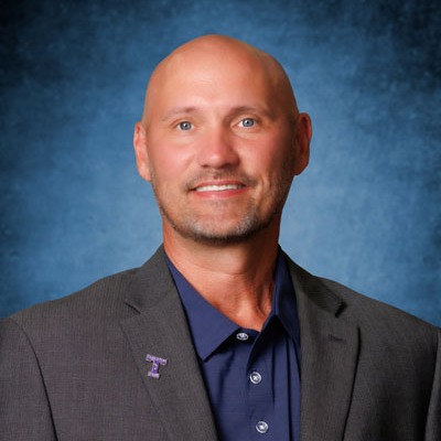 A man with a bald head smiles at the camera. He wears a gray jacket with a purple logo and a blue shirt.