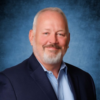 A man with white hair and a beard wearing a blue shirt and dark suit. He is smiling.