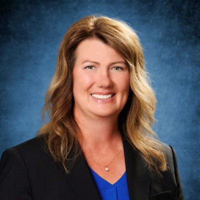 A woman with blonde hair smiles for the camera, wearing a blue shirt and a black jacket against a blue background.