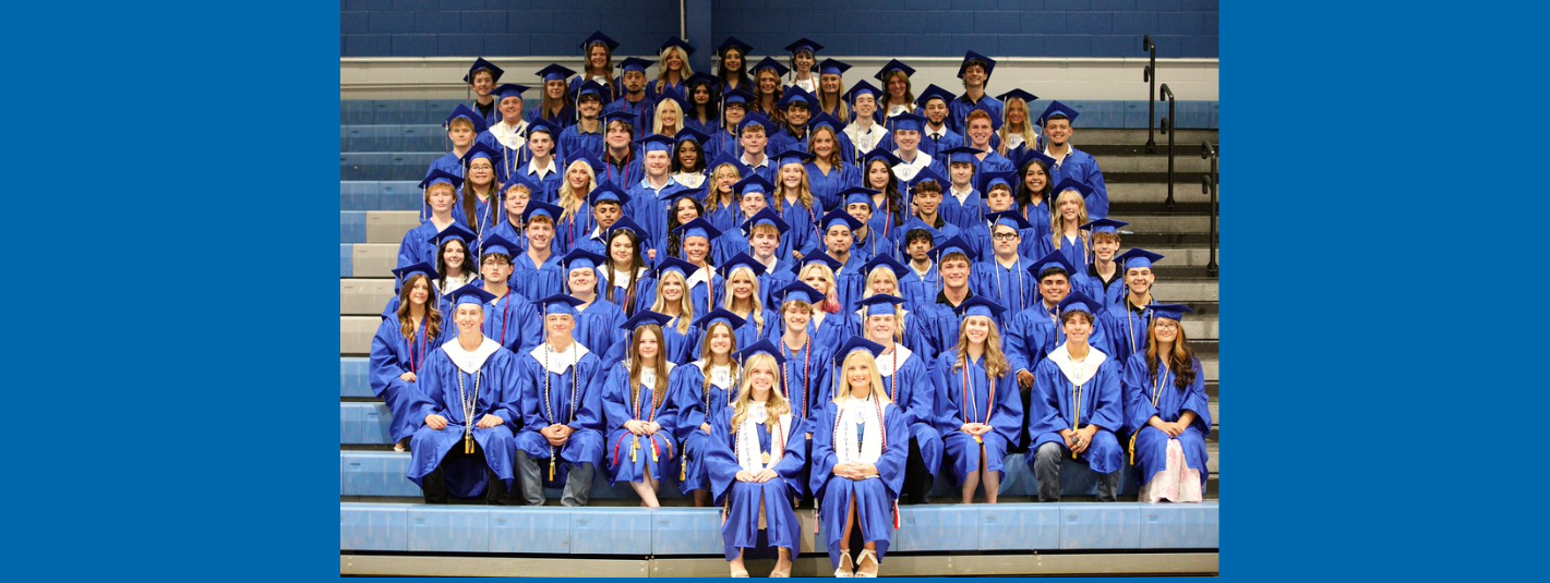 A group of graduates in blue gowns and caps pose for a photo on stadium bleachers.