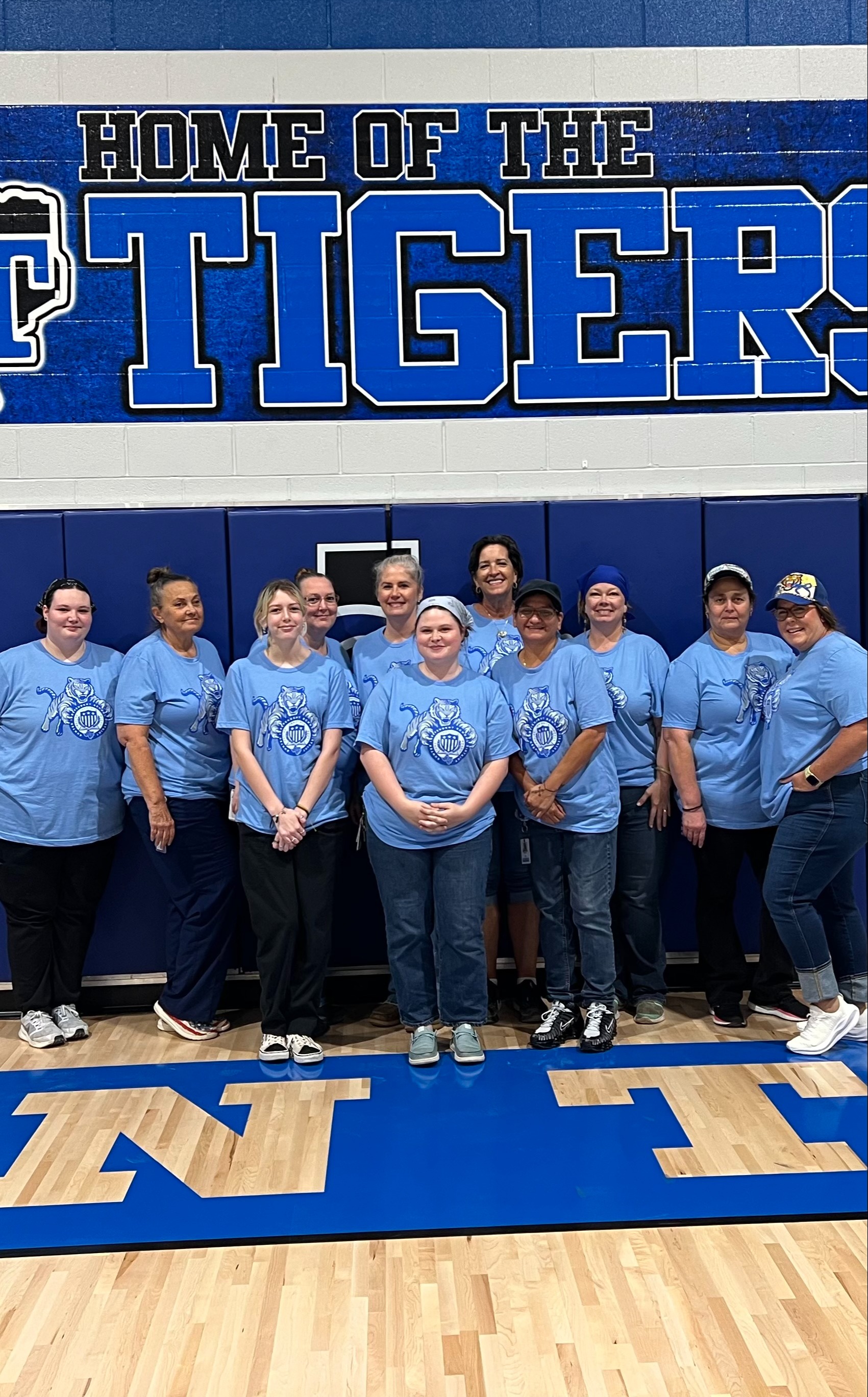 Group of people wearing blue shirts and jeans standing in a gym. A blue banner behind them reads "Home of the Tigers".