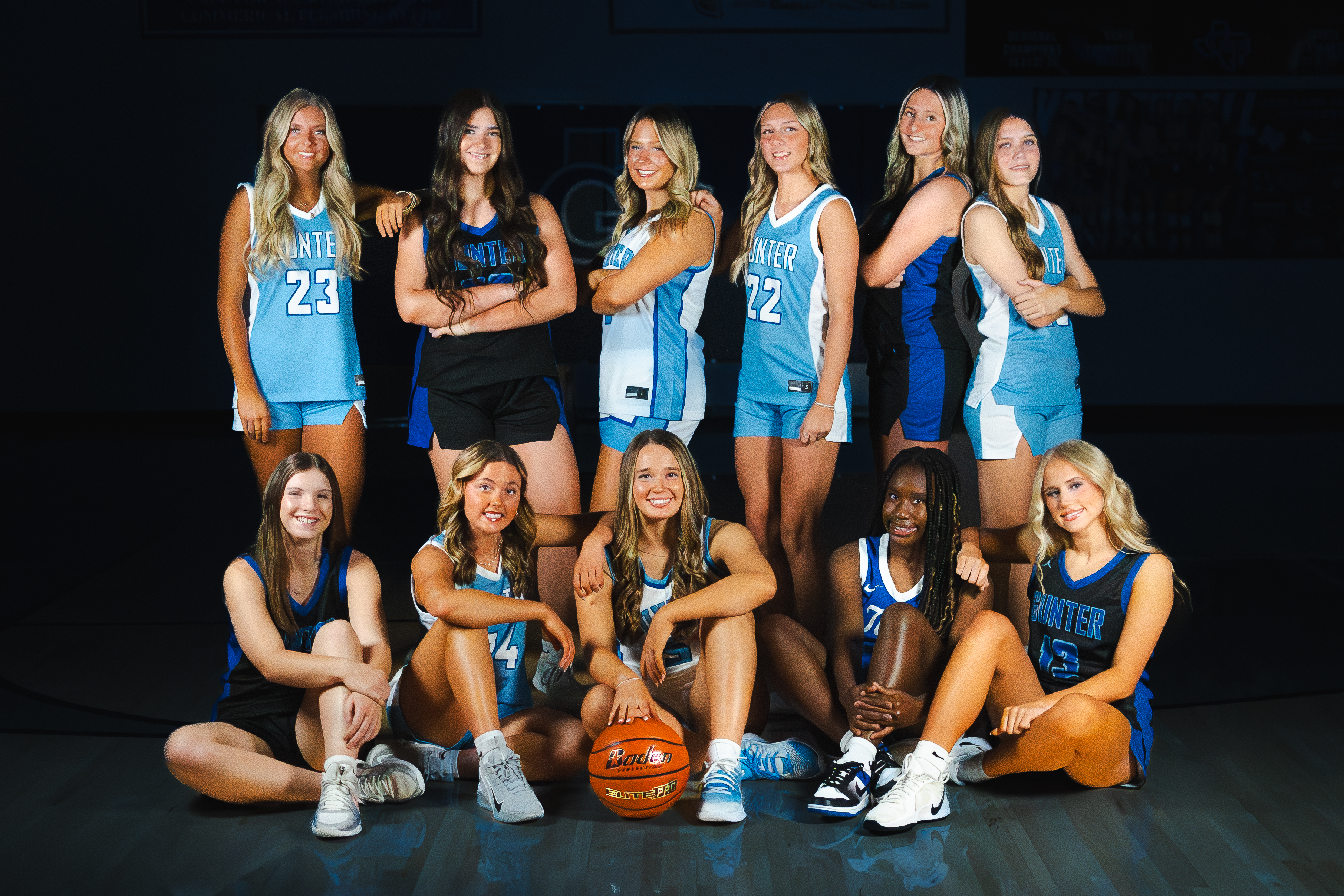 A group of girls in blue and white uniforms pose on a basketball court with a basketball.