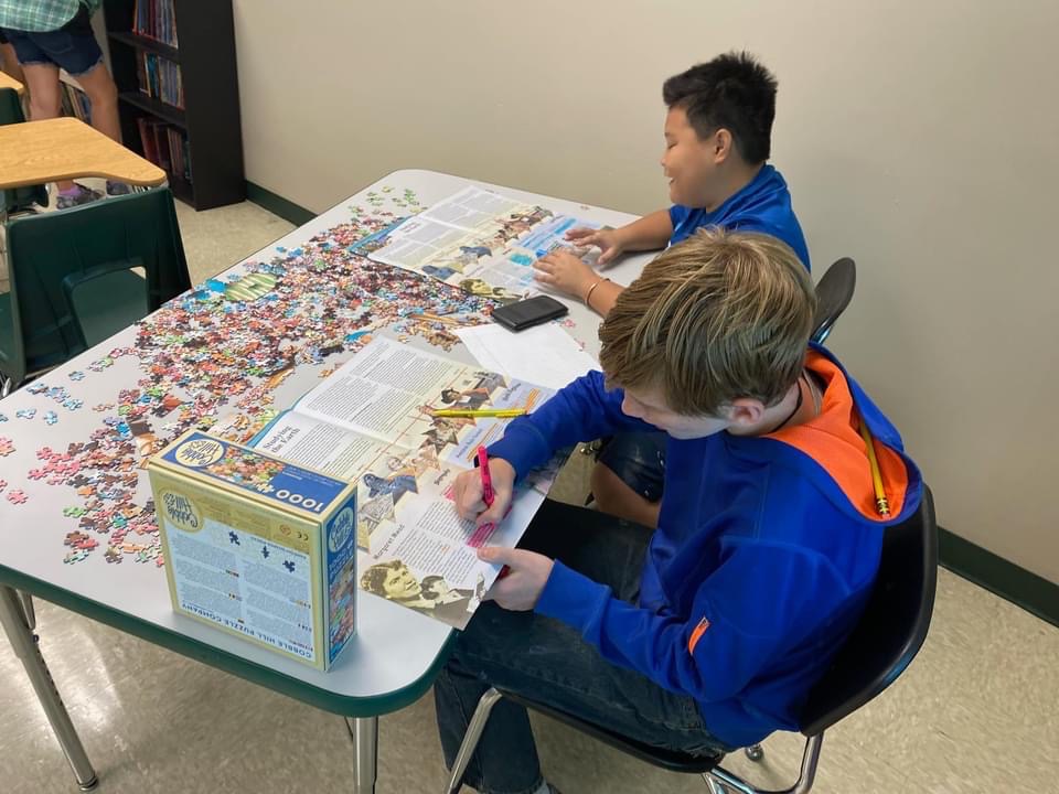 Two middle school boys working on newspaper with puzzle pieces on table.