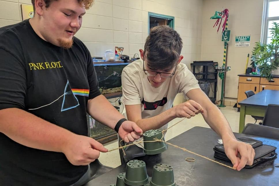 2 High school boys stacking cups using string and not hands.