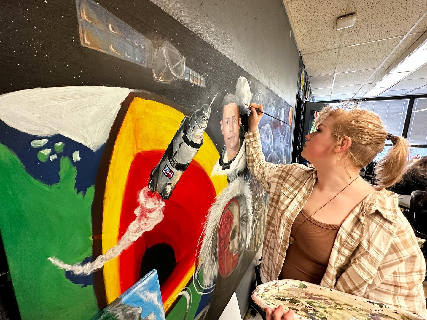 A student paints a large scale science mural at Marietta High School with  a cross section of the Earth, a rocket, Thomas Edison, an astronaut and a satellite. 
