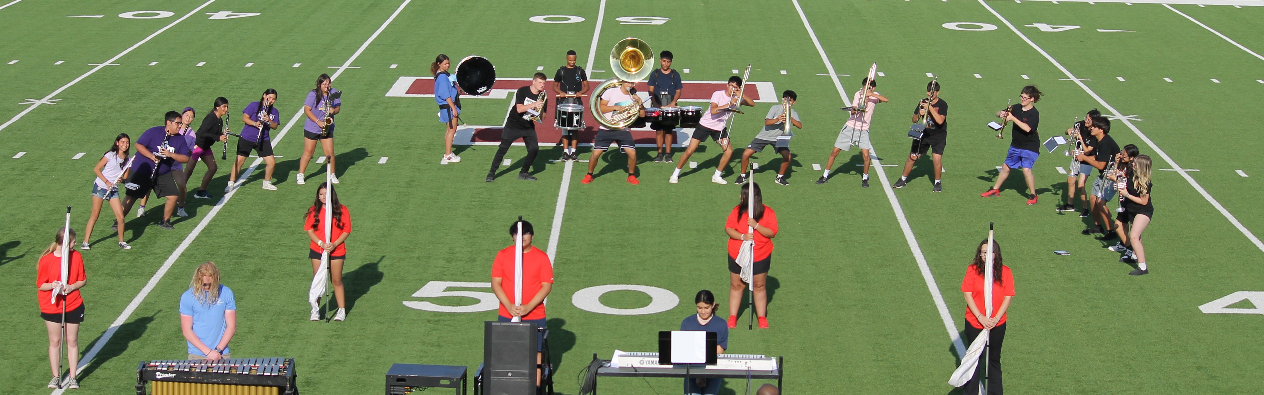 High-angle view of a high school marching band and color guard rehearsing on a green football field at the 50-yard line, viewed from the stands with instructors watching in the foreground.