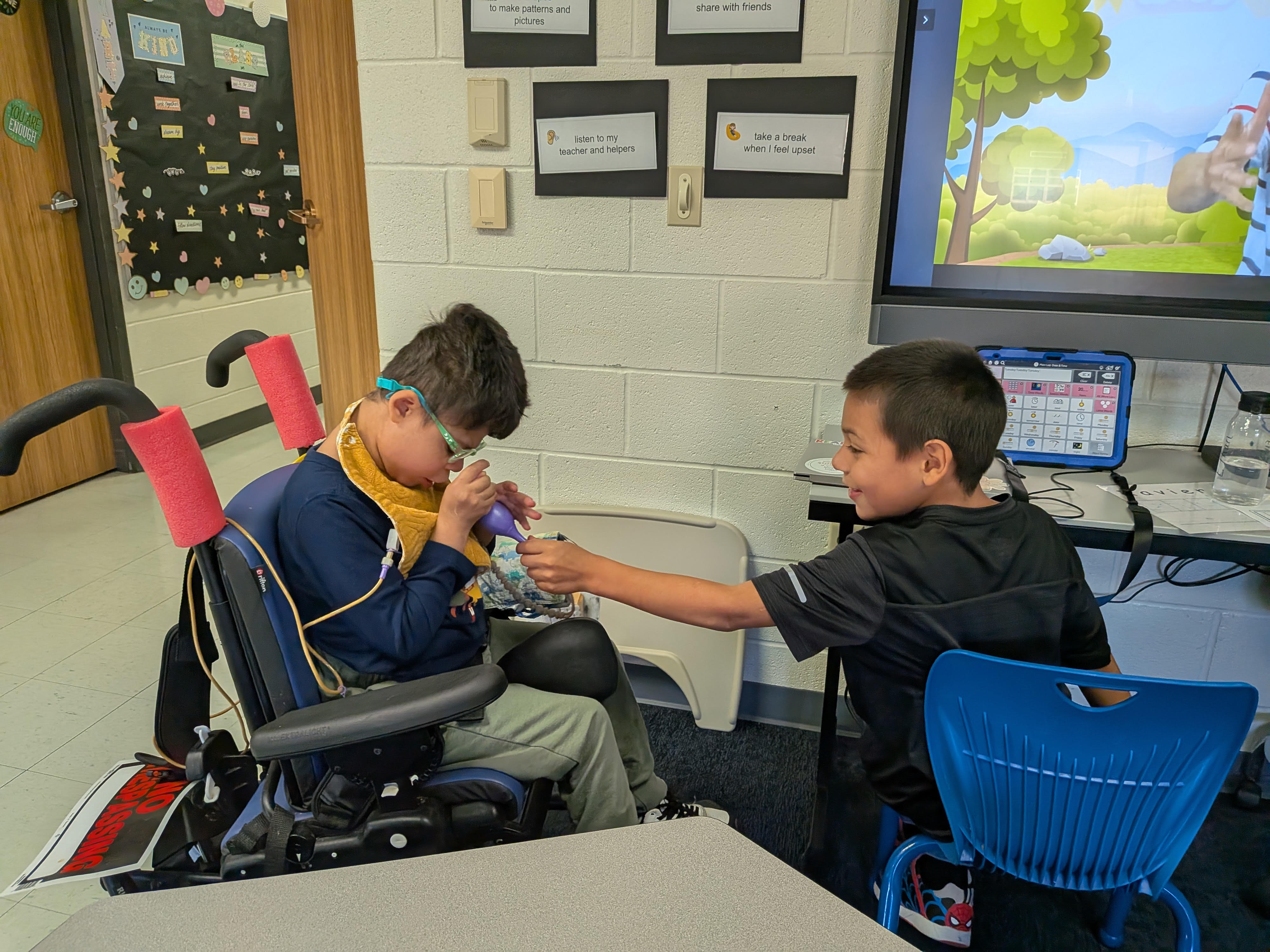 This image shows two children in a classroom setting. One child is seated in a wheelchair, wearing a yellow bib and glasses, and appears to be using a purple bulb syringe. The other child is sitting on a blue chair next to him, holding the other end of the syringe's tubing and smiling. In the background, there is a large screen displaying a cartoon scene and a tablet showing a grid of icons, possibly a communication app.