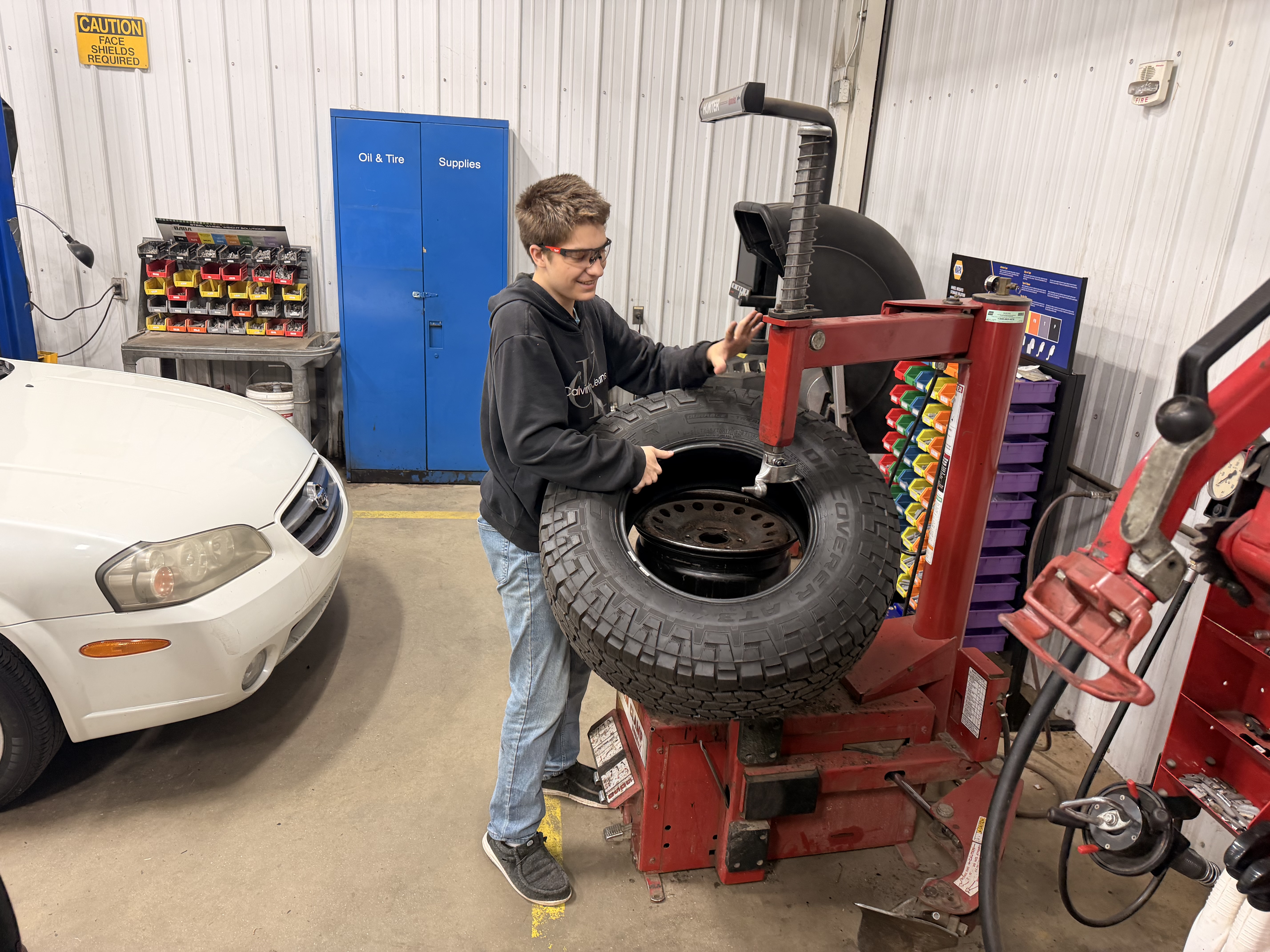 This image shows a young man operating a red tire changing machine in a workshop. He is wearing safety glasses, a black Calvin Klein hoodie, and blue jeans, and appears to be mounting or dismounting a large, heavy-tread tire from a black steel wheel. In the background, there are blue storage cabinets labeled "Oil &amp; Tire" and "Supplies," as well as a parts bin with colorful organizers.