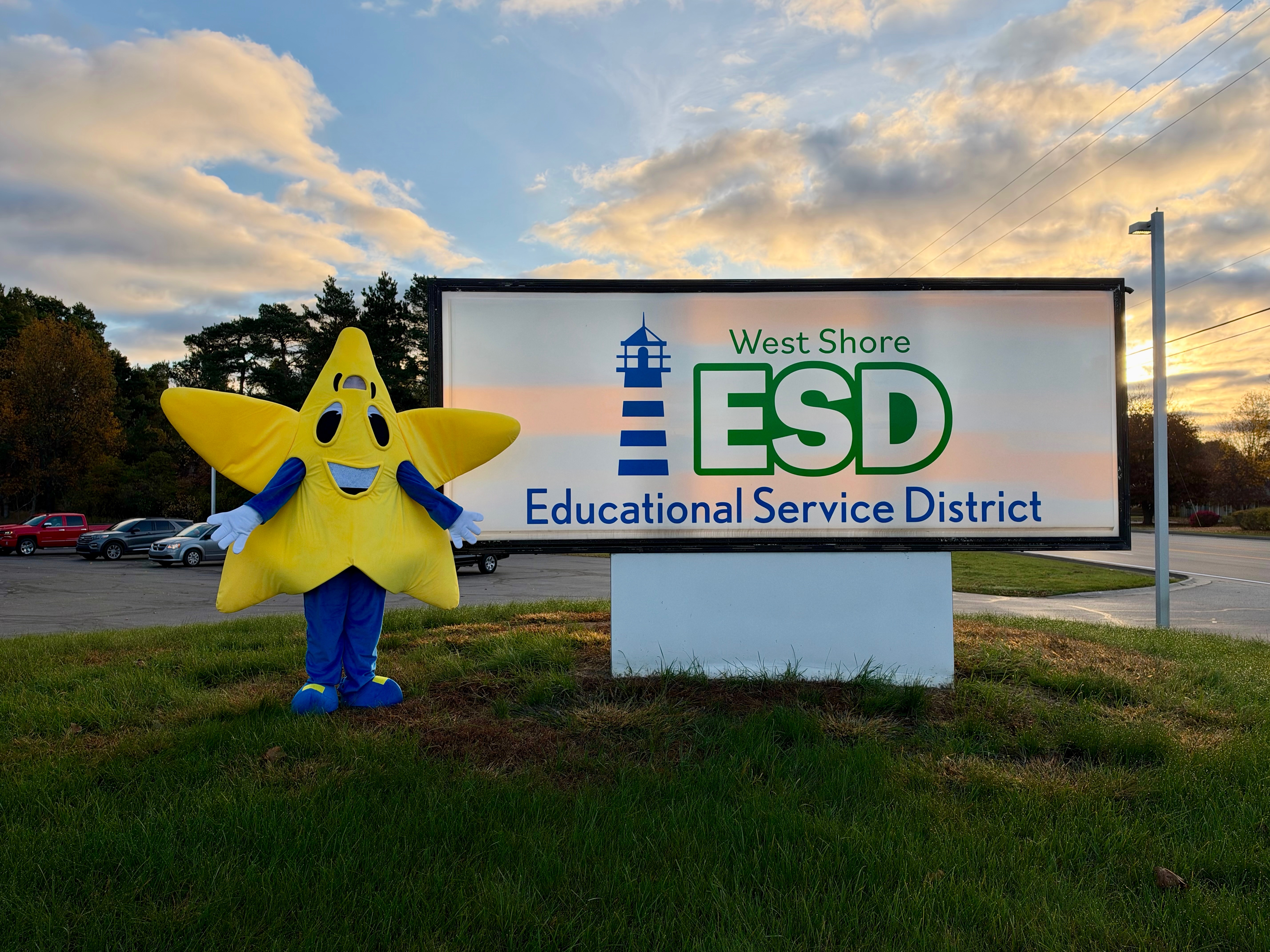This image features a large, yellow star-shaped mascot with blue arms and legs standing next to a sign for the "West Shore ESD Educational Service District." The sign also includes a blue and white lighthouse graphic. The mascot and sign are situated on a grassy area with a parking lot and trees in the background, under a cloudy sky at sunset.