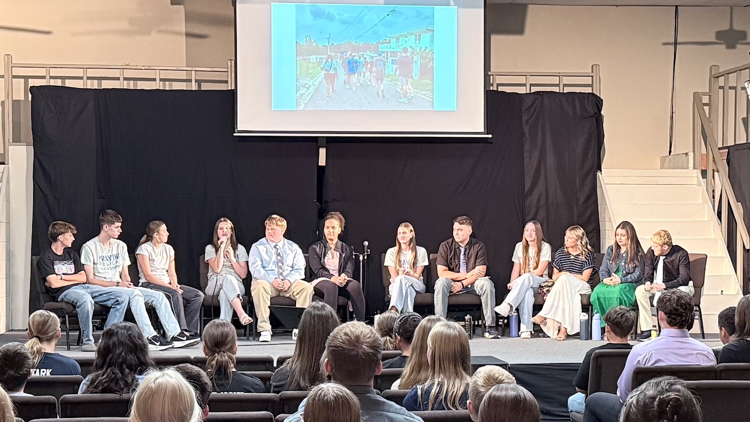 A classroom with students on stage, one speaking into a microphone. Behind them is a projector screen.