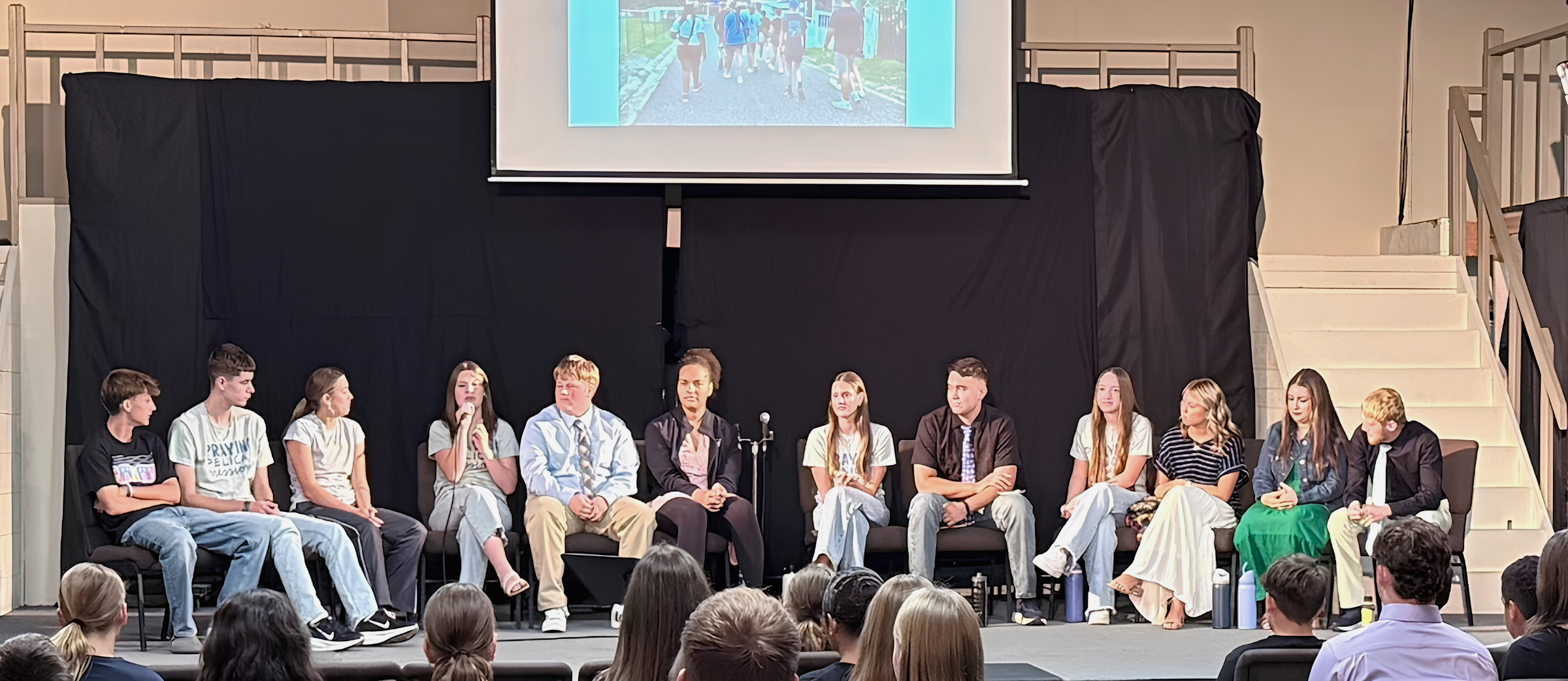 Panel of people on stage with a projector screen in the background and an audience in chairs.