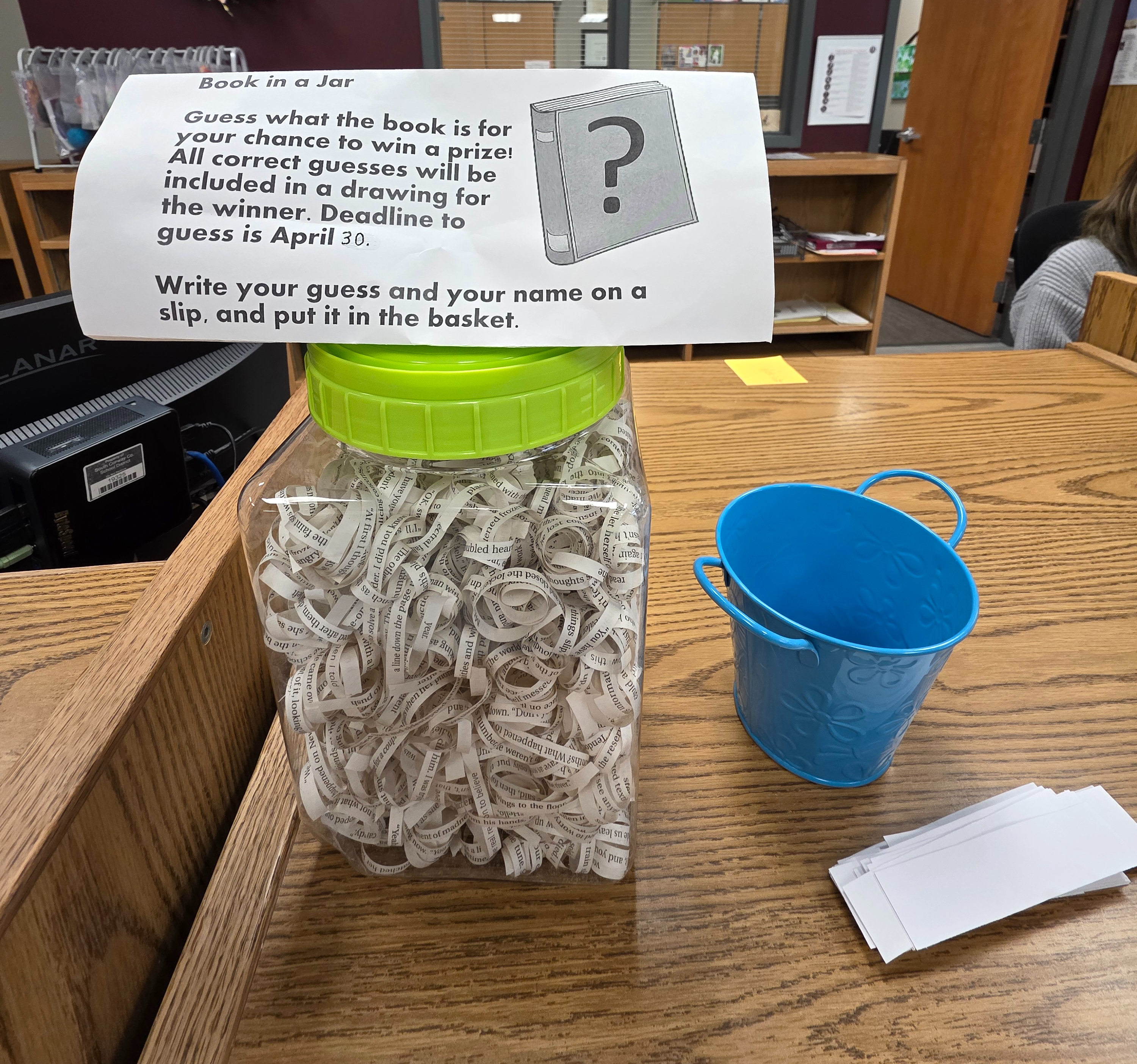 clear jar with green lid. inside jar is curled sentence strips from a book. Sign on top of jar announces Book-in-a-Jar contest. Blue canister beside it with white strips of paper in front for patrons to write their guess.