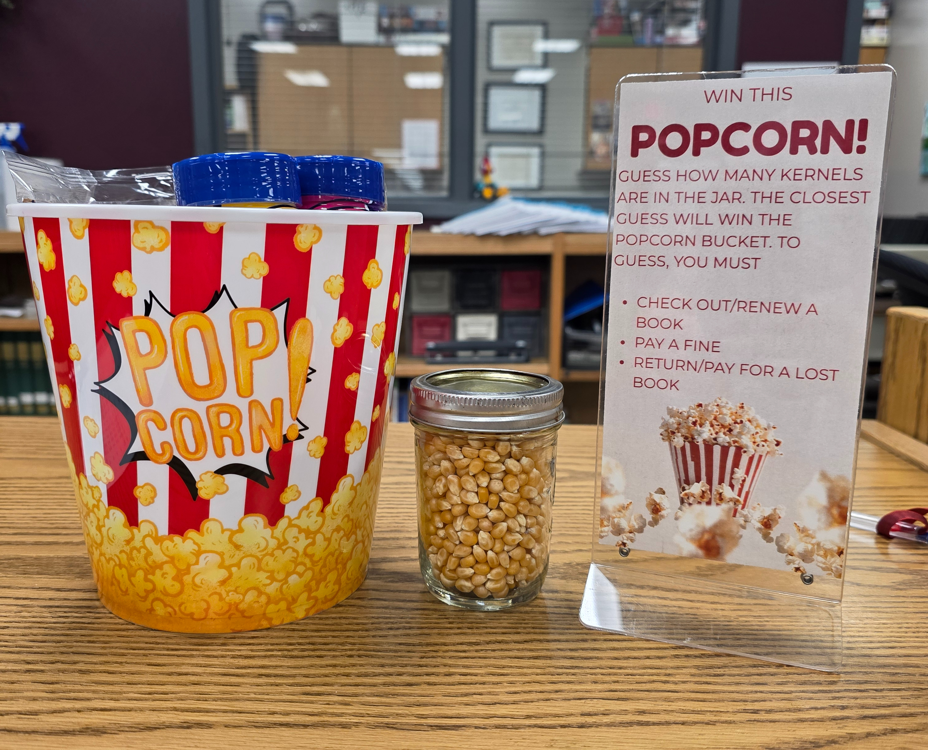 Red and white striped popcorn bucket filled with microwave popcorn packets, popcorn seasoning, and candy next to a clear jar filled with unpopped popcorn kernels and a sign announcing the contest rules