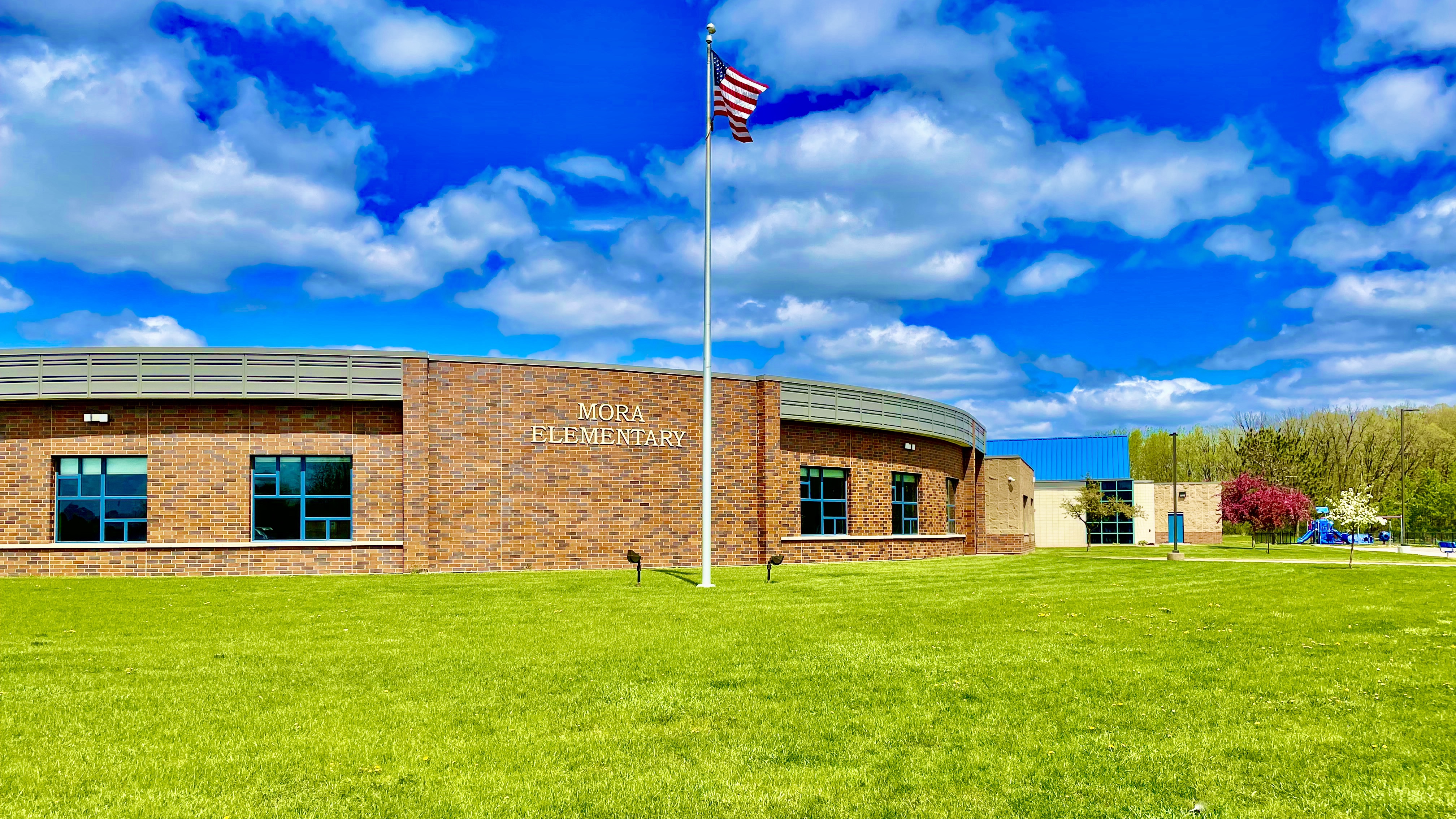 picture of school from outside, flag waving, green grass and bright blue sky