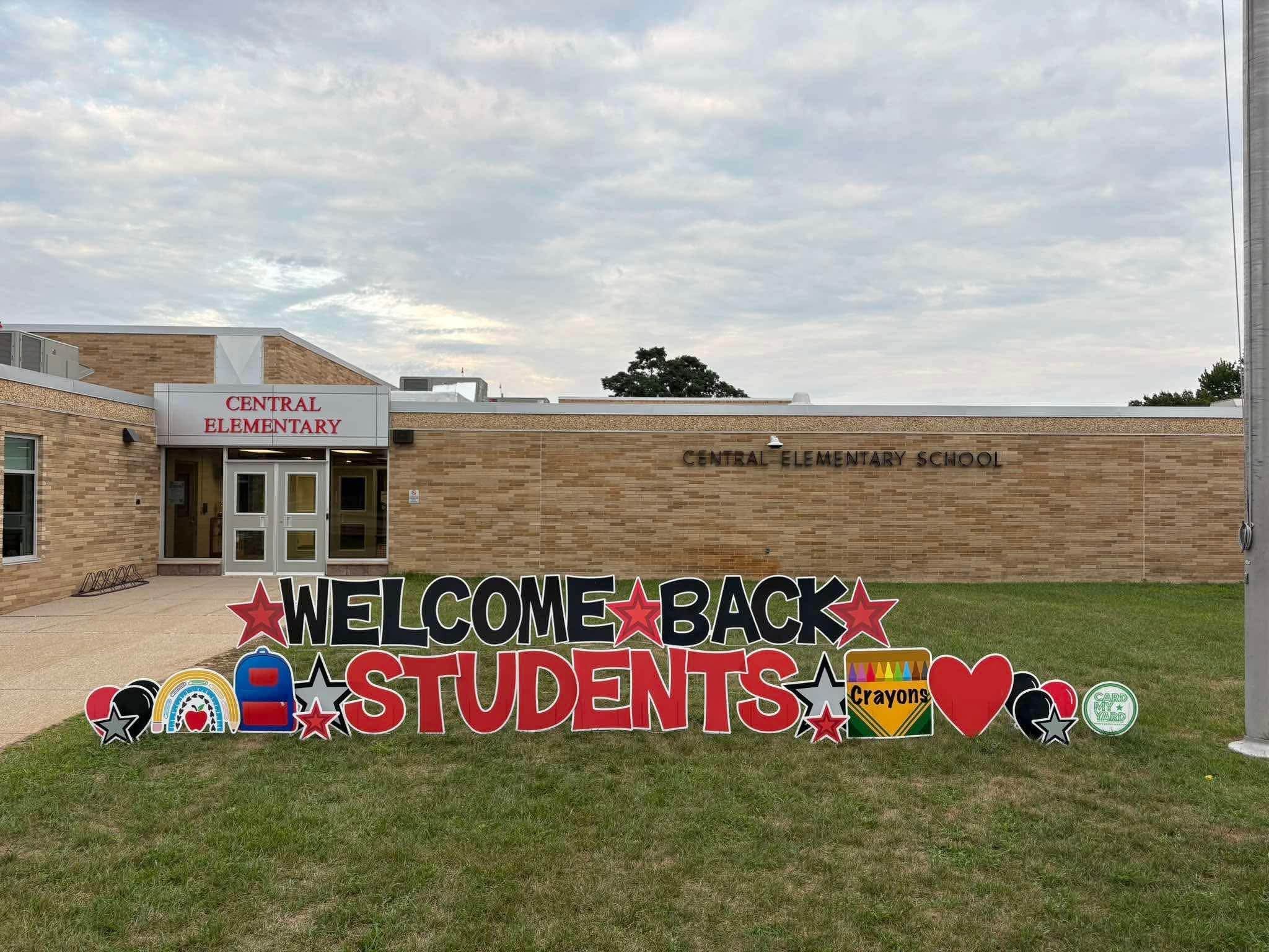 Welcome back sign with colorful colors, a box of crayons and a blue backpack. In front of the school building. 