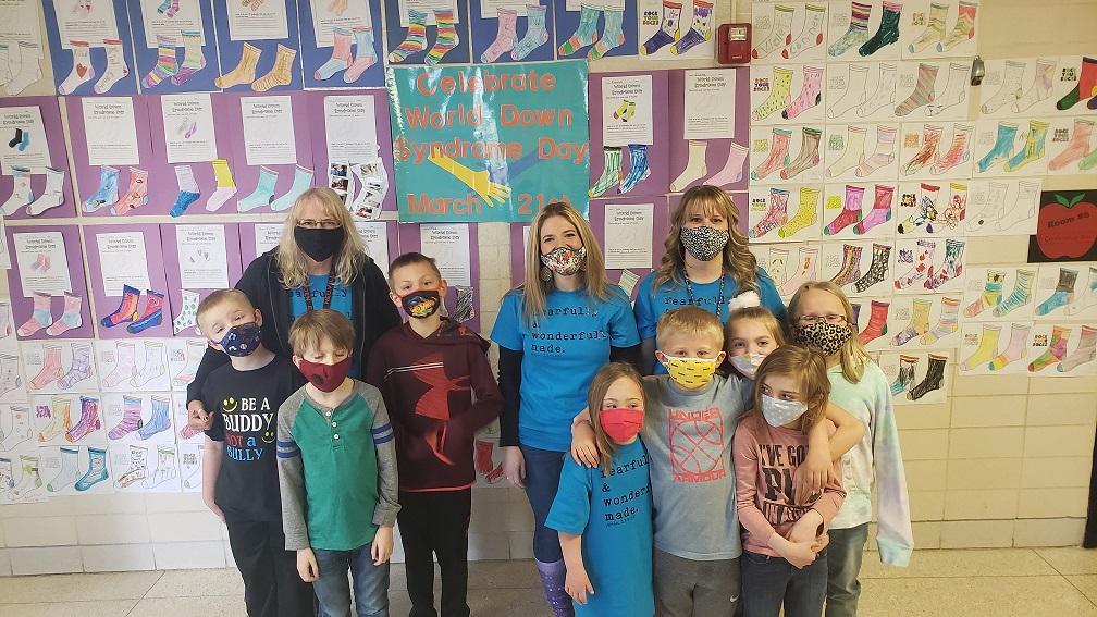 Several teachers and students wearing masks and standing in front of a bulletin board.