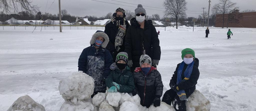 Students in winter coats and wearing masks behind a pile of large snowballs.